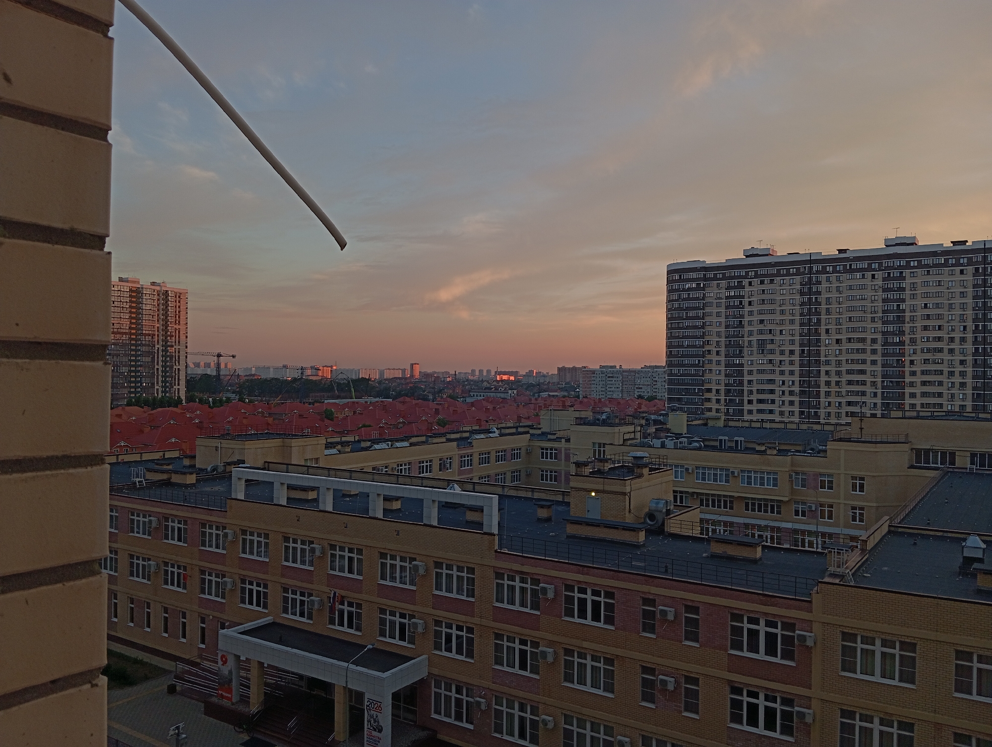 The image depicts a cityscape at dusk, likely in Krasnodar, Russia, given the architecture and style of buildings. The foreground is dominated by a section of a brick building, suggesting the photo was taken from a high vantage point. In the mid-ground, there are several educational institutions or office buildings constructed with a combination of yellow and pinkish bricks, featuring numerous windows and air conditioning units. The roofs are dark. Further in the background, a dense urban landscape unfolds with numerous residential buildings, including a large, multi-story apartment complex with many windows. The sky is a gradient of pale blue and orange, indicative of sunset or sunrise. A thin, white rod is angled across the upper left portion of the frame. Cranes are visible in the distance, suggesting ongoing construction. The scene is devoid of people, animals, or vehicles, presenting a quiet, urban vista at the end of the day.