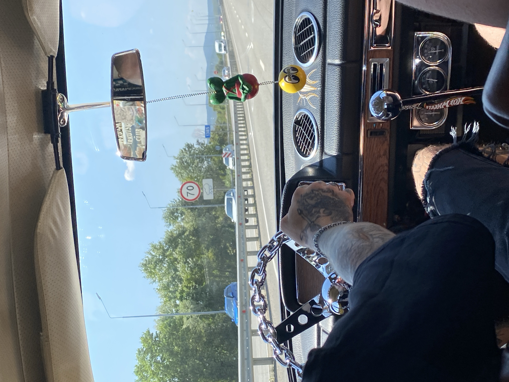 The image is taken from inside a car, looking forward through the windshield. The driver's hands are visible on the steering wheel, which has a large chrome chain around it. The interior of the car appears to be older, with wood-grain trim. A green alien-like air freshener and a yellow ball with a cartoon face hang from the rearview mirror. The car is on a highway, with other vehicles visible in the distance. Trees and greenery line the side of the road, and the sky is clear and blue. A speed limit sign indicating 70 is visible. The scene is consistent with a sunny day. The context suggests the car is traveling along the M4 Don Highway, a major federal highway in Russia, which passes through Goryachiy Klyuch, a resort city in Krasnodar Krai.
