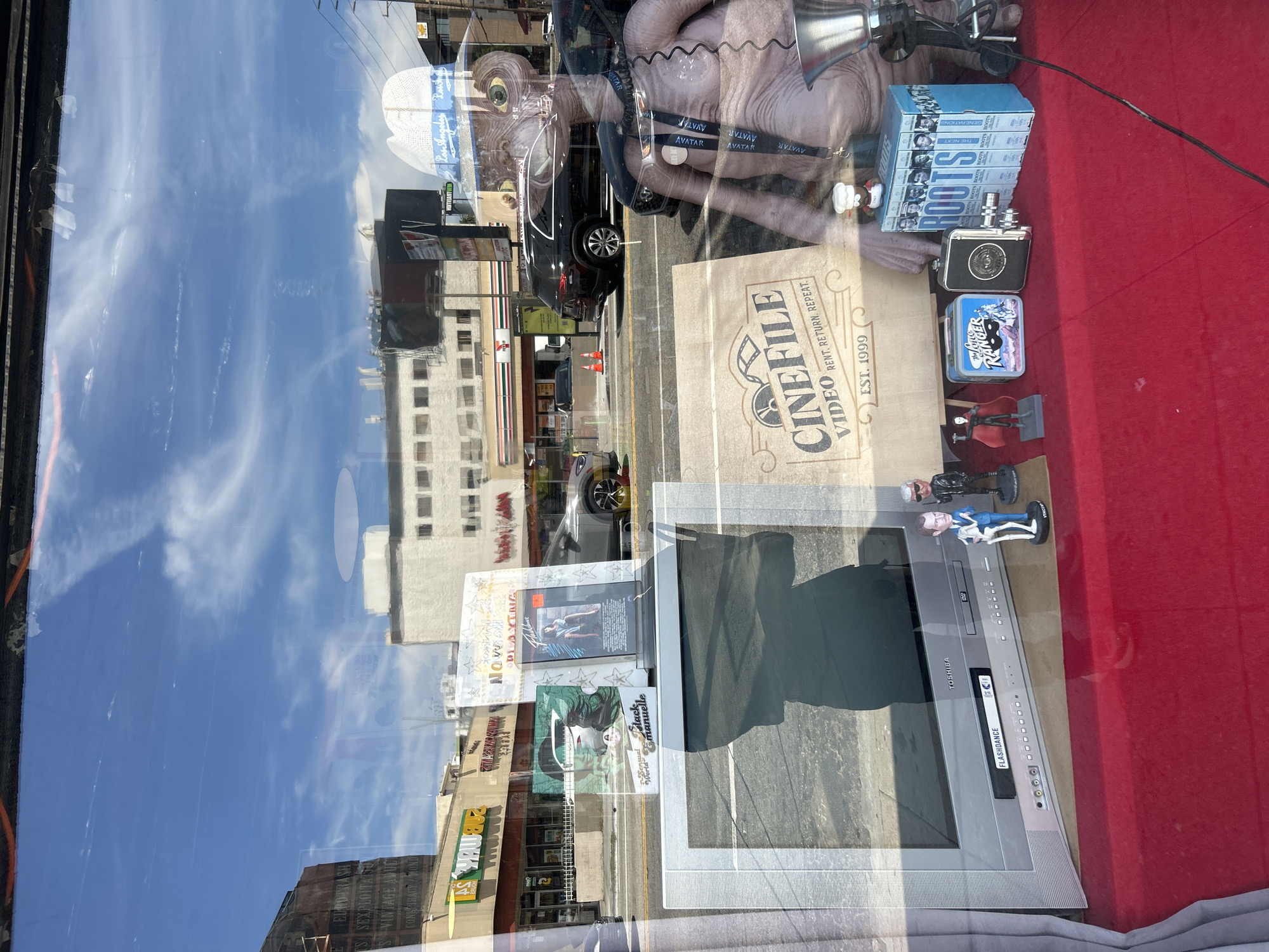 This image captures a window display in Los Angeles, United States of America. The display includes a Cinefile Video bag, which suggests the location is near a Cinefile video store. Other items visible are VHS tapes, figurines, and a Toshiba TV/VCR combo. The window reflects the street outside, including buildings, a Subway restaurant, and the sky. An alien figurine wearing a hat is also part of the display.