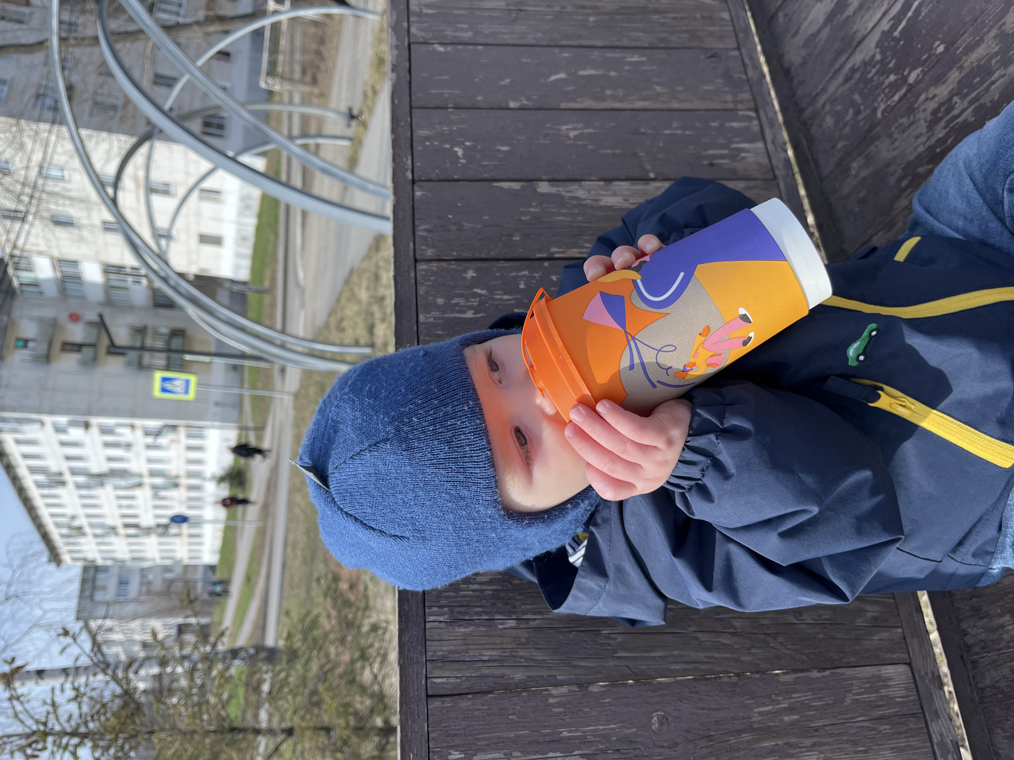 A child sits on a wooden bench outdoors, holding a colorful cup to their face. The child is wearing a blue knit hat and a navy blue jacket with yellow trim. In the background, there is a metal structure, a street, and a building with multiple windows. A street sign is visible near the building. The location is Chaikovsky, Russia.