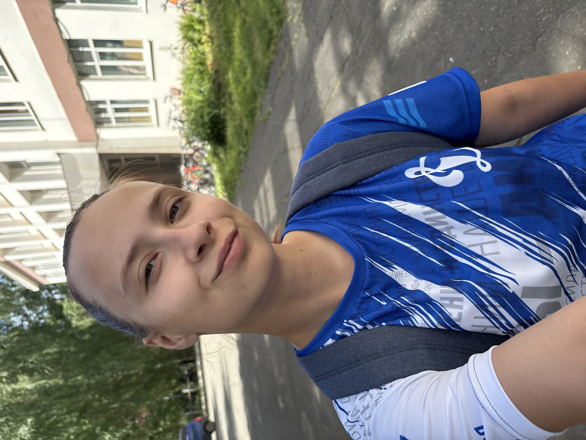 In a selfie taken outdoors in Chaikovsky, Russia, a young Caucasian female with her hair pulled back is visible from the chest up, wearing a blue and white athletic t-shirt with a gray backpack. She is smiling slightly and looking towards the camera. The background includes a building with windows and a red-brown band, a sidewalk, green grass, and several parked bicycles. A portion of a blue car is also visible on the left side of the image. The lighting suggests it is daytime, likely sunny. The scene appears to be in front of a school or similar institution.
