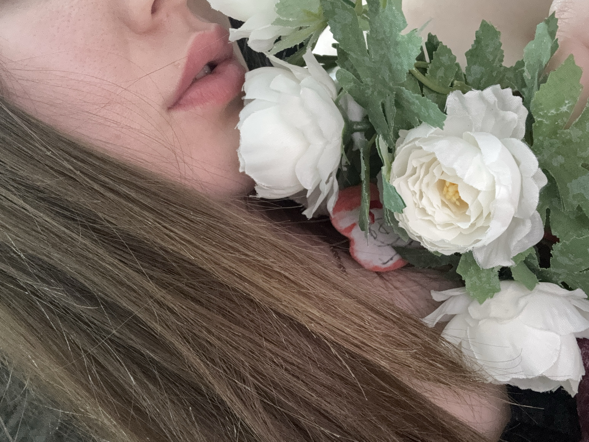 A close-up shot shows a portion of a person's face, including lips and cheek, adorned with white flowers and green leaves. Long, brown hair is also visible. The image does not contain enough information to determine a specific location in Chaikovsky, Russia.