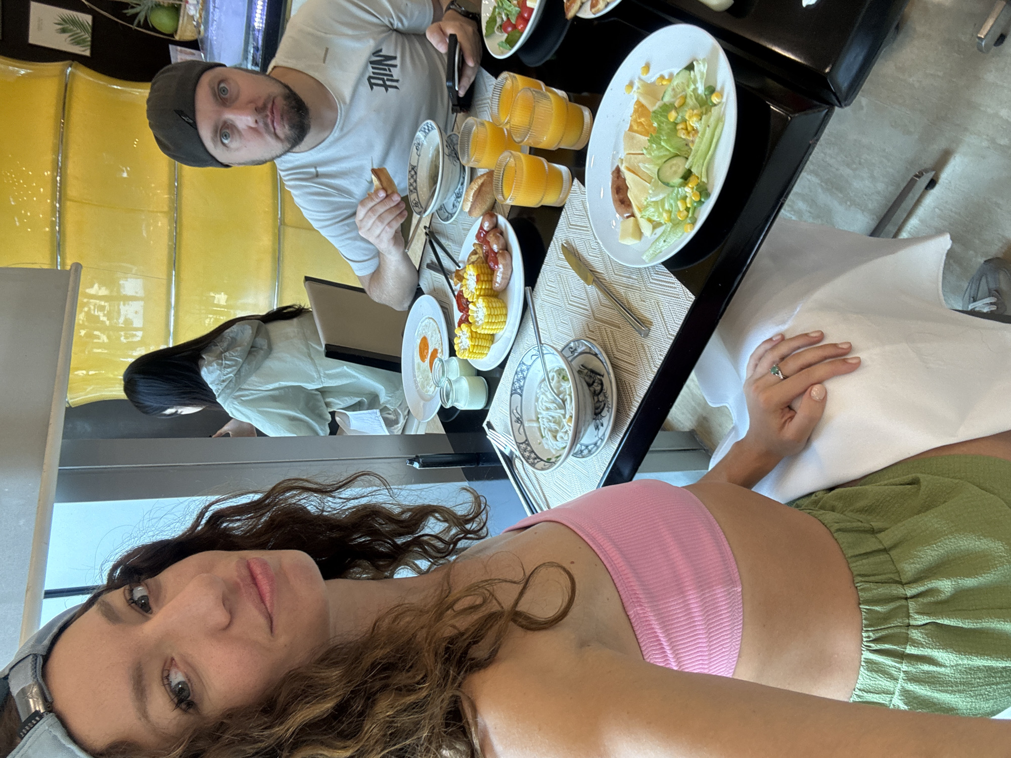 The image shows a selfie taken at a restaurant in Đà Nẵng, Vietnam. A woman in a pink top and green shorts is holding the camera. Behind her at the table is a man in a white t-shirt and grey beanie. The table is laden with breakfast foods including salad, eggs, corn, sausages, bread, and orange juice. Other people are also sitting at the table. The interior of the restaurant includes a yellow decorative wall.