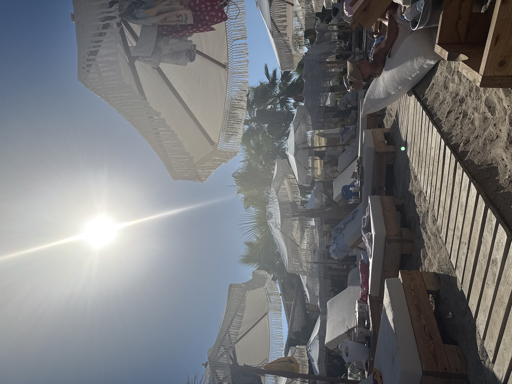 The image depicts a sunny day at a beach resort, likely in Pallini Municipal Unit, Greece. The scene is oriented vertically, showing a clear blue sky with a bright sun and lens flare dominating the upper left. Below the sky, several white beach umbrellas with decorative fringes are visible, some open and others stacked. Palm trees can be seen peeking through the umbrellas, suggesting a warm, tropical or Mediterranean climate.
In the foreground and midground, there are wooden beach loungers with white cushions, arranged on a sandy beach. A wooden walkway is laid out on the sand, providing access to the loungers. Some people are visible relaxing on the loungers. One person, wearing a straw hat, is lying on a lounger, holding what appears to be a phone. Another person is partially visible sitting on a lounger. Scattered items like towels and water bottles are present on the loungers and the sand.
The overall impression is one of leisure and relaxation in a picturesque beach setting. The strong sunlight indicates it is daytime, likely midday. The presence of beach umbrellas, loungers, and palm trees are strong indicators of a resort or a well-equipped beach. No explicit text is clearly legible, though some patterned fabrics on the umbrellas might contain faint designs or logos. The mood conveyed is tranquil and summery.