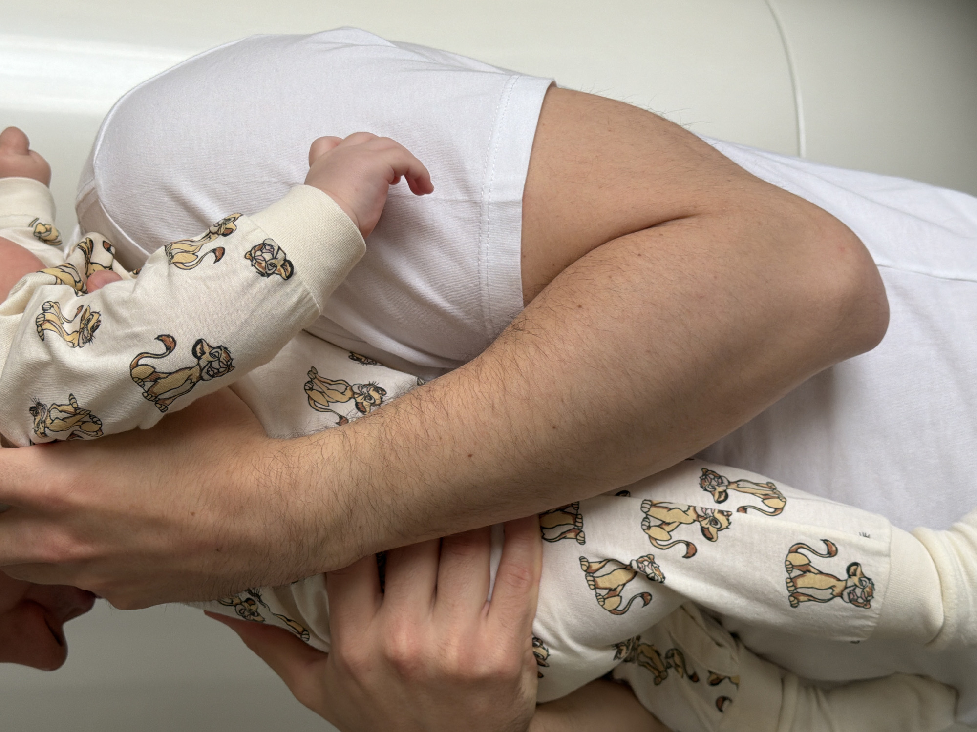 The image captures a warm, intimate moment between an adult and a baby, set indoors, likely in Khimki, Russia. The scene focuses on an adult, wearing a simple white t-shirt, holding or cuddling a baby. The baby is dressed in cream-colored pajamas or a onesie adorned with a repeating pattern of cartoon lion cub characters, identifiable as Simba from Disney's The Lion King.
The baby's small hands and arms are visible, reaching towards or around the adult, suggesting a tender embrace. The adult's arm, showing some hair, is also visible around the baby, reinforcing the sense of closeness and protection. The background consists of soft, light-colored fabric, possibly a bed or a couch, bathed in bright, diffused lighting, indicating an indoor setting, likely during the daytime. The overall impression is one of comfort, love, and security shared between the two individuals. No specific text is visible in the image apart from the character patterns on the baby's clothing.