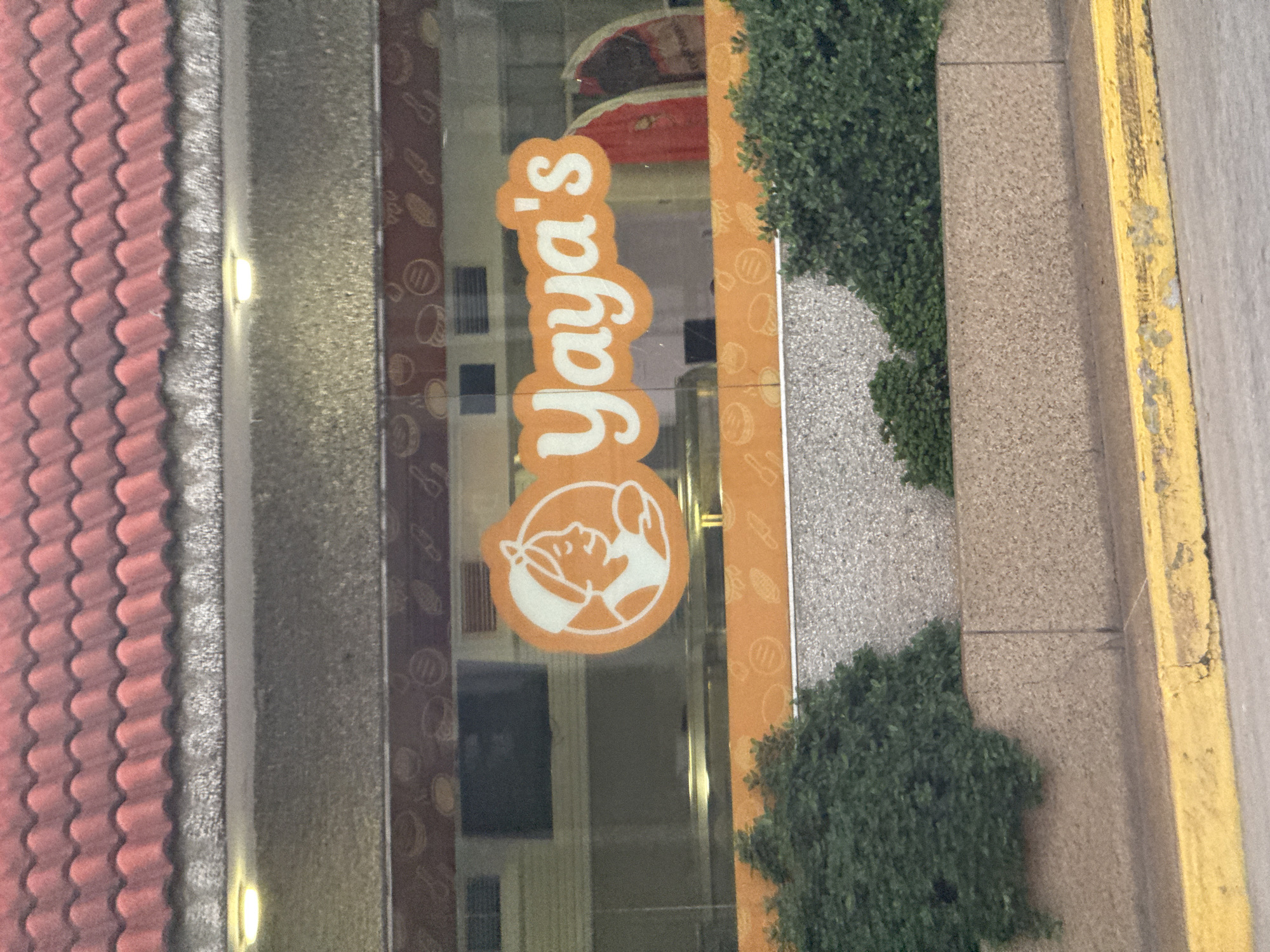 The image shows the exterior of a restaurant in Los Robles, Venezuela. The restaurant, named "Yaya's", has a prominent orange and white logo on its storefront window. The logo features a stylized depiction of a woman holding a baby, enclosed in a circle. The name "Yaya's" is written in white, cursive lettering against an orange, cloud-like shape. The storefront also features decorative bands with repeating circular patterns and food-related icons such as bread and forks.
The building has a red tile roof and a concrete facade. In front of the storefront, there are well-maintained green bushes and a textured concrete wall. A yellow curb and asphalt road are visible in the foreground, suggesting a street-level view. The lighting suggests it is daytime, with one visible light fixture illuminating the exterior. The overall impression is that of a clean and established eatery in a commercial or public area.