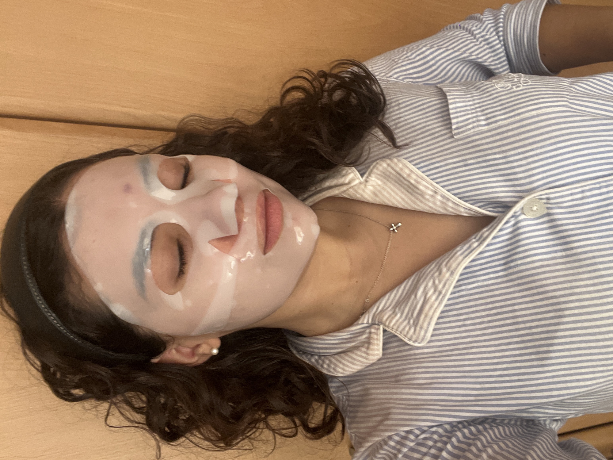 A woman with dark curly hair is lying on a wood surface, wearing a white and blue striped collared shirt and a sheet face mask. She is also wearing a thin black headband and a delicate silver necklace with a small pendant. The location is unknown and cannot be determined from the image.
