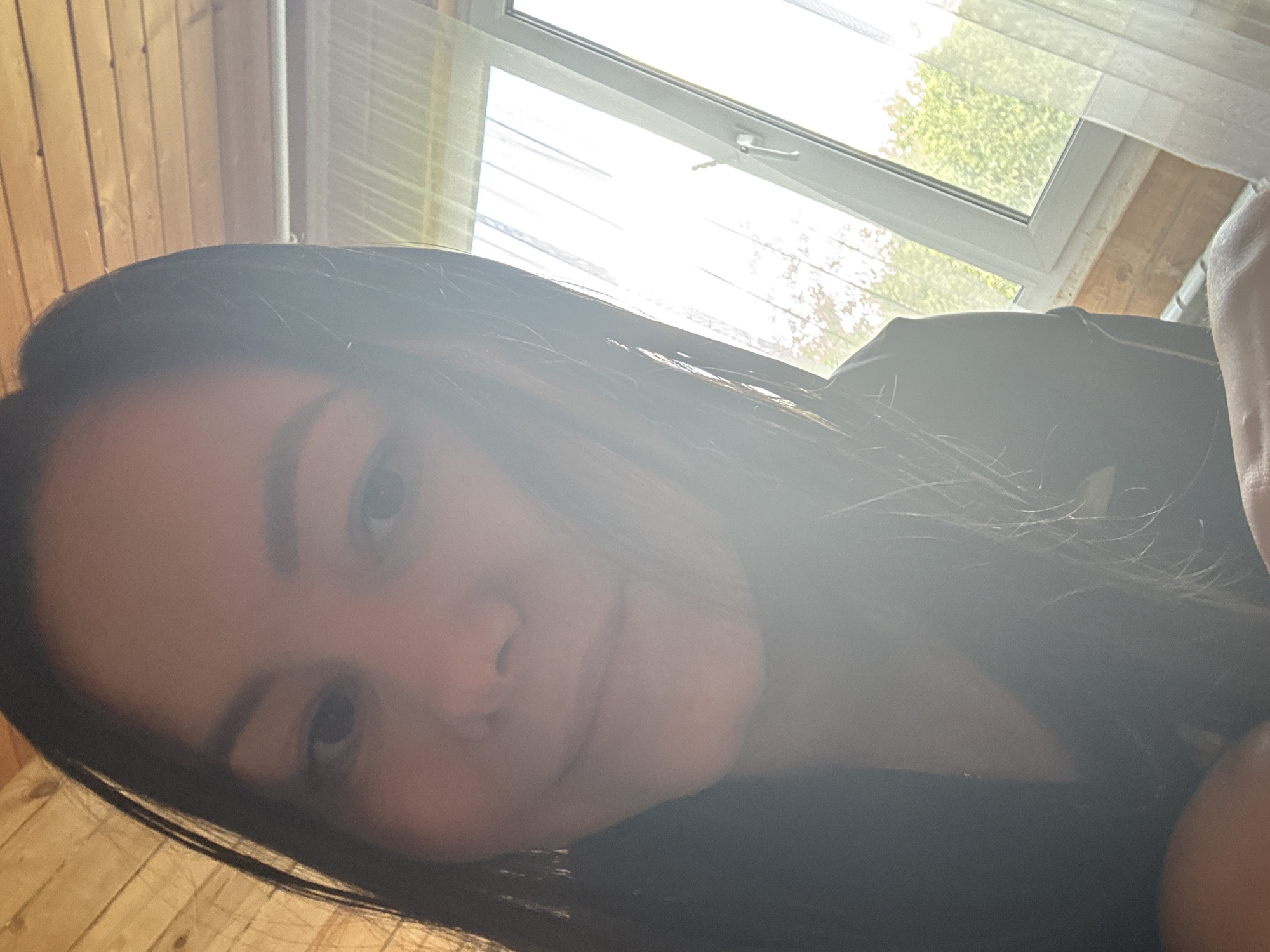 A medium shot, indoor selfie of a young woman with long dark hair. She is looking directly at the camera with a soft smile. Her face is illuminated by natural light coming from a large window to her right. The window is partially covered by sheer white curtains and shows green foliage and a bright sky outside. The room appears to be a loft or attic space, with wooden plank walls and ceiling. The woman is wearing a dark-colored garment. The overall lighting is bright, with some lens flare obscuring parts of the image. The location is Nikolaevka, Russia.