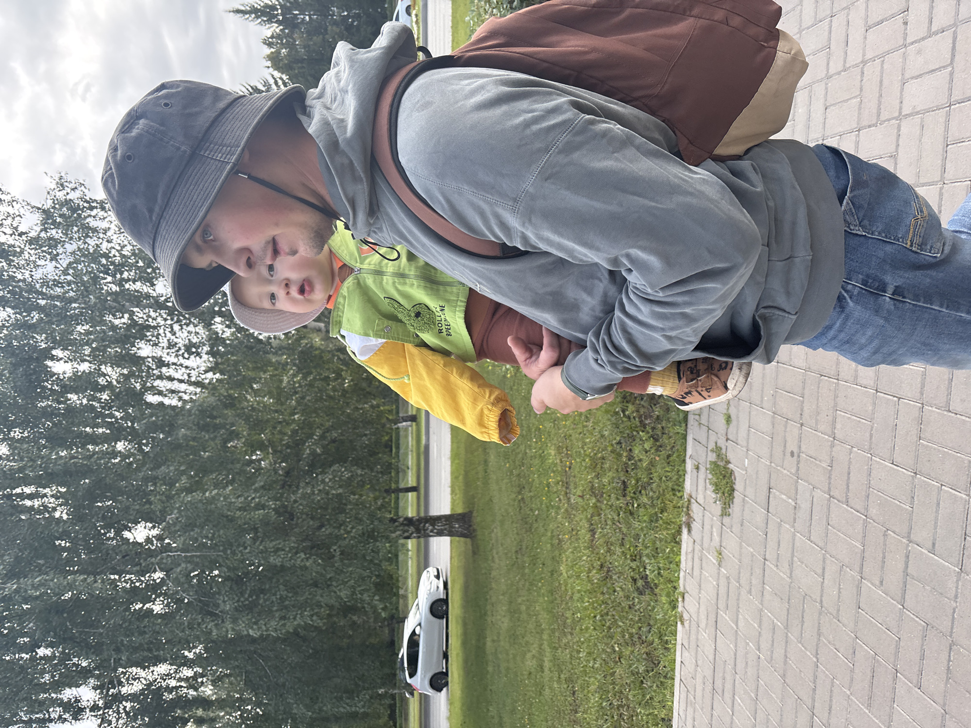 A man wearing a bucket hat, a grey hoodie, and blue jeans carries a young child dressed in a yellow and green jacket and a pink hat. The man is walking on a paved sidewalk next to a grassy area with trees in the background. A white car is visible driving on a road in the distance. The scene appears to be an outdoor urban environment in Naberezhnye Chelny, Russia. The man is looking to the right, while the child is looking forward with a surprised expression. A brown backpack is slung over the man's right shoulder.