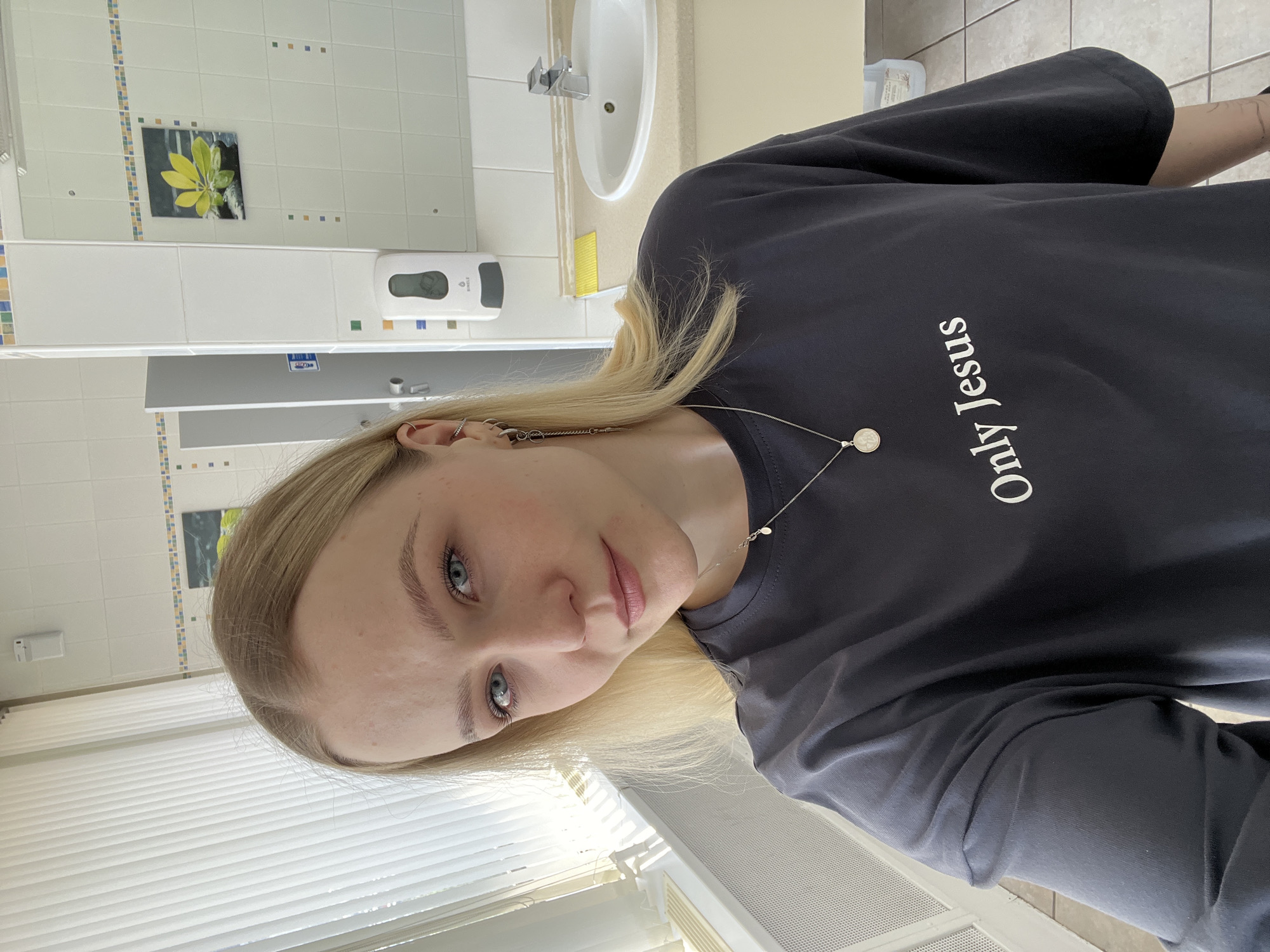 A young woman is taking a selfie in what appears to be a public restroom. She has blond hair, blue eyes, and is wearing a dark-colored t-shirt with the words "Only Jesus" printed on it. She also wears a necklace with a small pendant.
The background features white tiled walls with a decorative border, a mirror, a sink, a soap dispenser, and a painting of a leaf. A window with white blinds is visible in the lower left corner. The floor has a gray, tiled surface.
There is no information available in the image to confirm that the location is in the city of Khimki, Russia.