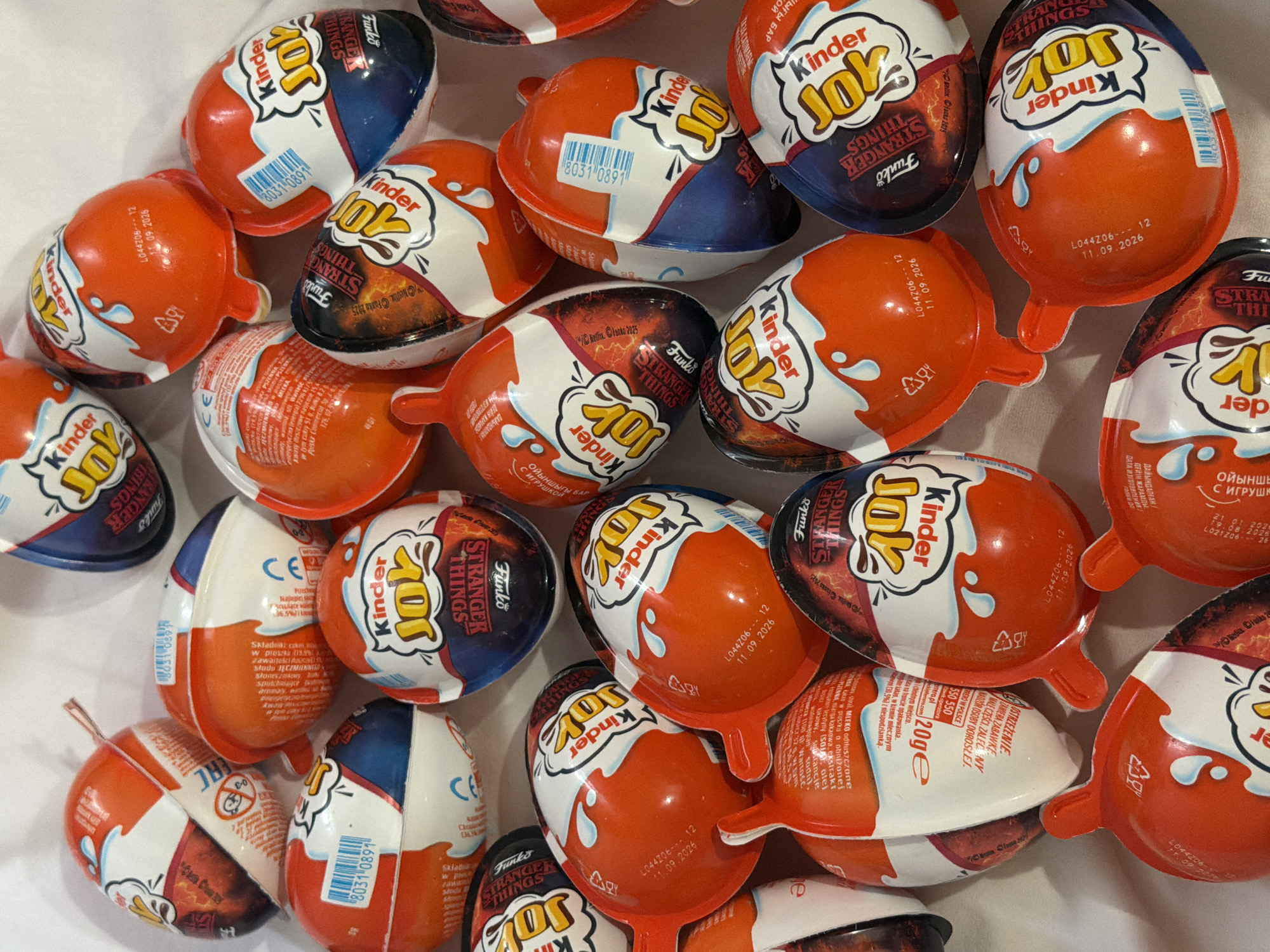 A close-up photograph shows a pile of Kinder Joy chocolate eggs. The eggs have a split design with one half being orange and the other half being white or blue, and all have the Kinder Joy logo. Some are marked with "Funko Stranger Things," which indicates a collaboration with the TV series Stranger Things. There are production codes and expiration dates printed on the packaging. The setting suggests they are on a white surface, possibly a table or countertop. The location is not discernible from the image.