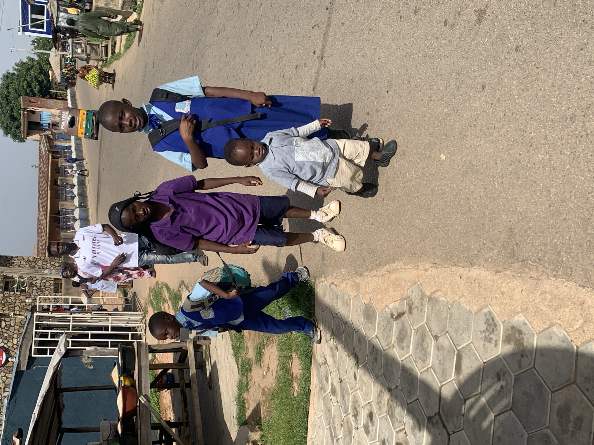 The media file displays a group of people, primarily children, walking on a street in what appears to be a populated area in Jos, Nigeria. The primary subjects are several children, some of whom are wearing school uniforms. One child in the foreground is wearing a blue and white school uniform with a blue tie and a backpack. Another child next to them is wearing a purple shirt and dark shorts. A younger child, possibly a toddler, is also present, dressed in a light grey polo shirt and light-colored shorts. There are other individuals in the background, including adults and more children.
The scene depicts a street with an asphalt surface, bordered by a tiled walkway on one side and buildings and vegetation on the other. The architecture suggests a marketplace or a residential area with small shops or homes. A rickshaw-like vehicle, common in some parts of Nigeria, is visible in the background. The time of day appears to be daytime, with clear shadows cast by the people and objects, indicating sunny weather.
The children seem to be either going to or returning from school, suggested by their uniforms and backpacks. Their expressions are neutral, with the child in the foreground looking towards the camera. The overall atmosphere seems to be that of daily life in a bustling urban or semi-urban environment.
Visible text includes "Emirates Fly Better" on a white t-shirt worn by an adult in the background. The license plate on the rickshaw-like vehicle has "PTA 1696" written on it. There are also some indistinct logos on the school uniforms.