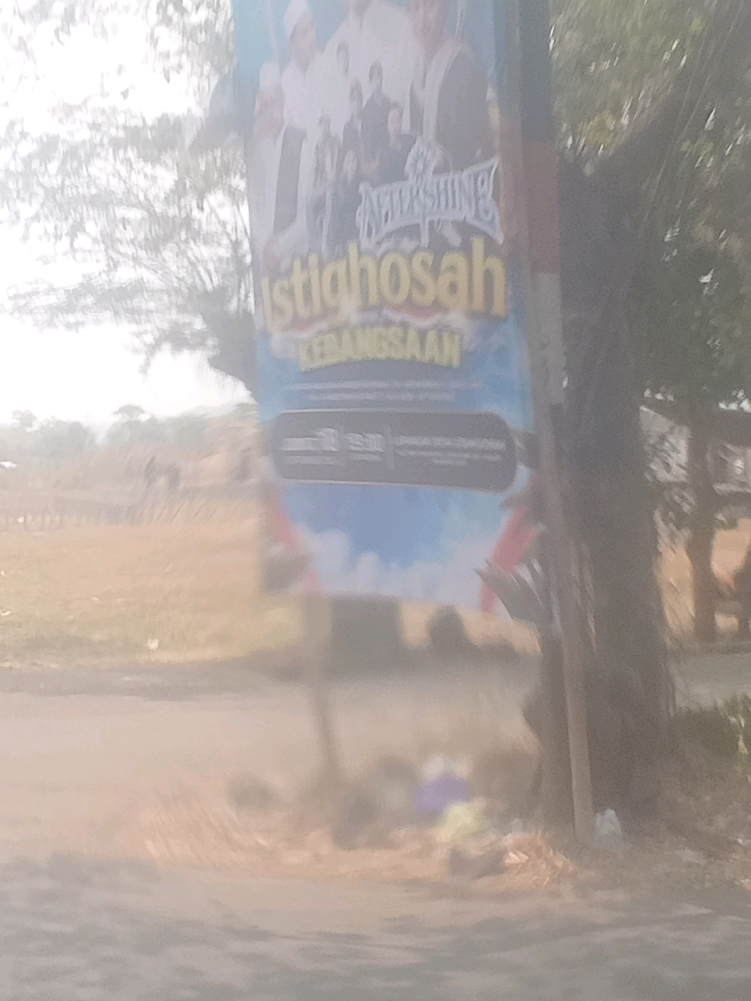 The image depicts a faded banner on a pole, likely in Lebakgowah, Indonesia, given the text on the banner. The banner has a blue background and features an image of several people, possibly a religious gathering or event. Text on the banner reads "Istiqhosah" and "KEBANGSAAN" in yellow lettering. Below this, there is a dark rectangular area with faint text, possibly containing event details. The banner is slightly obscured by a tree trunk to its right. The background shows a dry, grassy area with some vegetation and trees. The overall image quality is poor, with significant blur and overexposure, making it difficult to discern finer details.