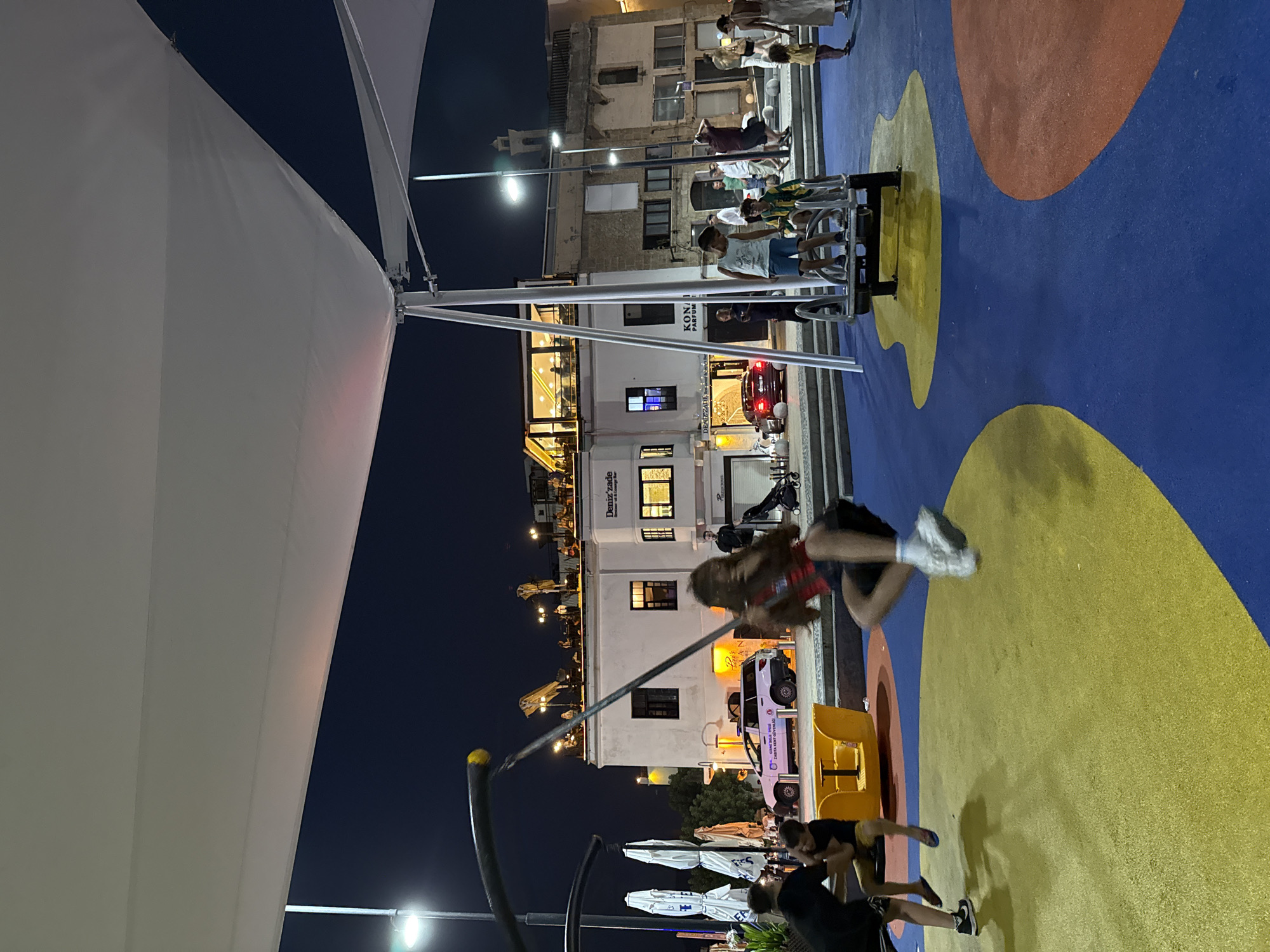 In an outdoor public space at night, several children are engaged in play. One child is actively swinging on a swing set, captured mid-air with their legs extended. Another child is seated on a rotating playground ride, holding onto a bar. In the background, a white building with multiple windows and a rooftop terrace is illuminated, with people visible on the terrace. Adjacent to the building, a pink vehicle with markings is parked, and several other individuals are standing or walking around the area. The ground surface is a colorful playground with distinct sections of blue, yellow, and orange rubberized material. Streetlights cast a glow over the scene, and the sky is dark.