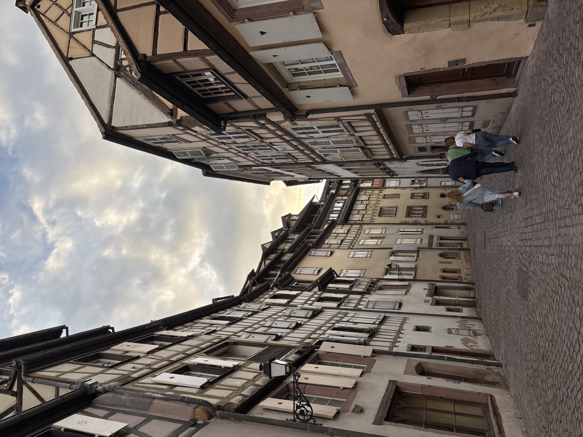 The image captures a picturesque cobblestone street in Colmar, France, lined with traditional Alsatian half-timbered houses. The architecture features distinct wooden beams against pastel-colored facades, with many windows adorned with white shutters, some intricately carved with heart shapes. A group of four people, two men and two women, are walking away from the camera down the street. The sky above is overcast with dramatic clouds, suggesting either dawn or dusk, casting a soft, diffused light on the scene. The perspective draws the viewer down the narrow, curving street, emphasizing the charming and historic atmosphere of the location, likely within the Petite Venise (Little Venice) district, known for its well-preserved medieval and Renaissance buildings.
