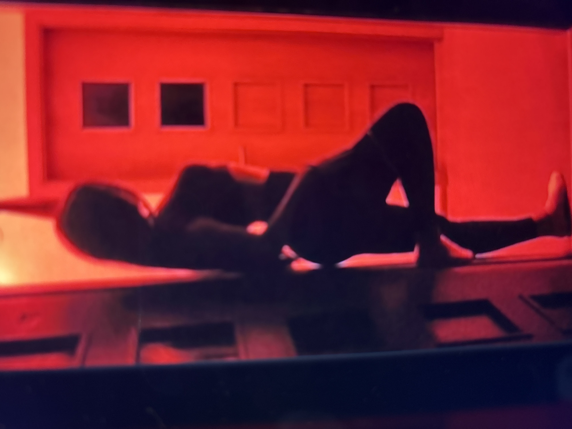 The image shows a silhouette of a person leaning against a dark surface on the left, with their back to the viewer. The person is wearing dark clothing, possibly athletic wear. To the right is a bright red door with two square inset panels, one above the other. The overall lighting is predominantly red, casting a dramatic glow on the scene. The background appears to be an indoor setting, possibly a hallway or entryway, with the red door being the focal point.