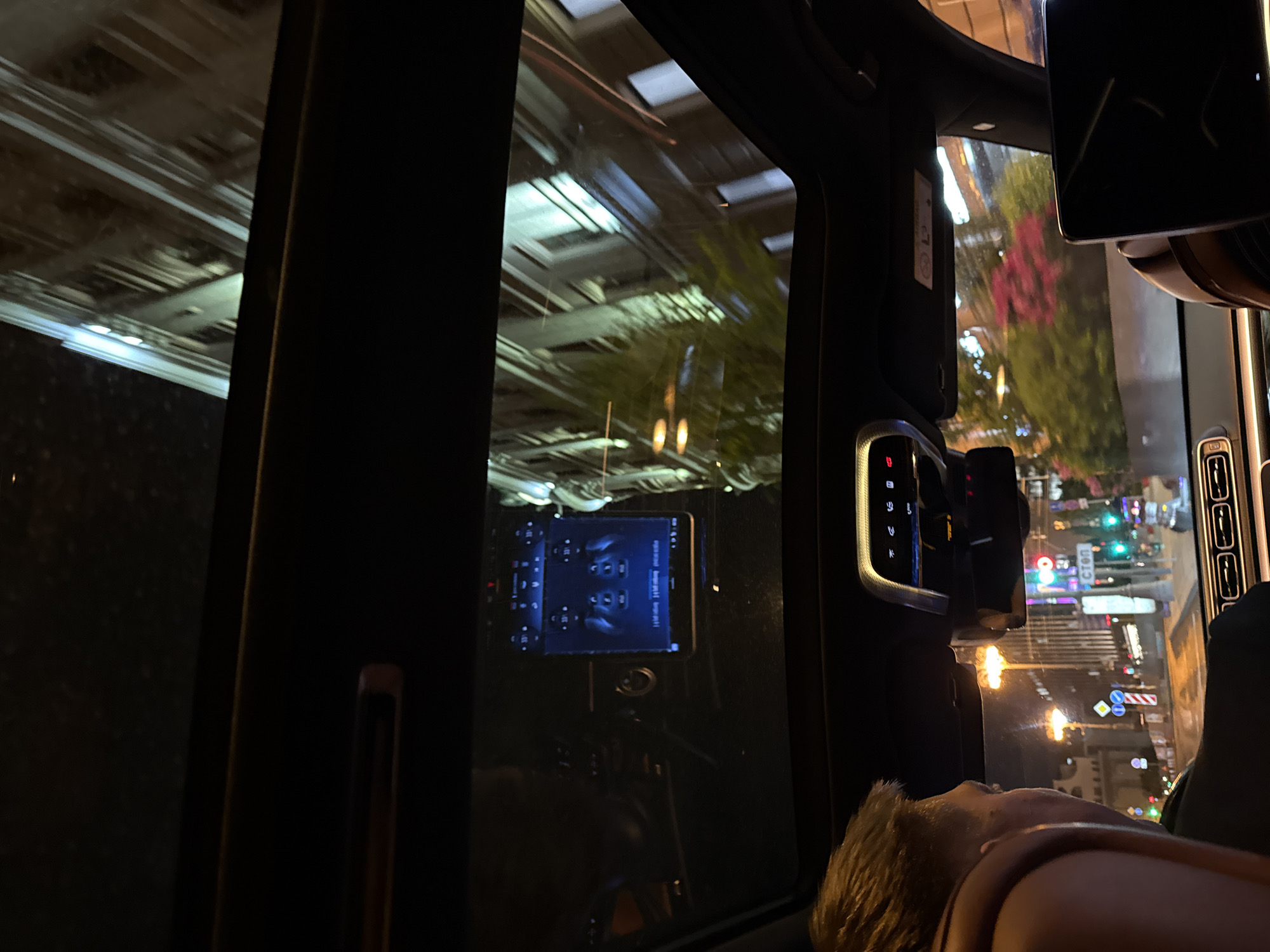 A nighttime shot taken from inside a vehicle moving through Moscow, Russia. The cityscape is visible through the side window, with blurred lights and signs indicating urban activity. Inside the car, the dashboard with control buttons and a screen are visible. A person’s head and shoulder are in the foreground, suggesting they are a passenger. Buildings and streetlights are seen through the side windows.