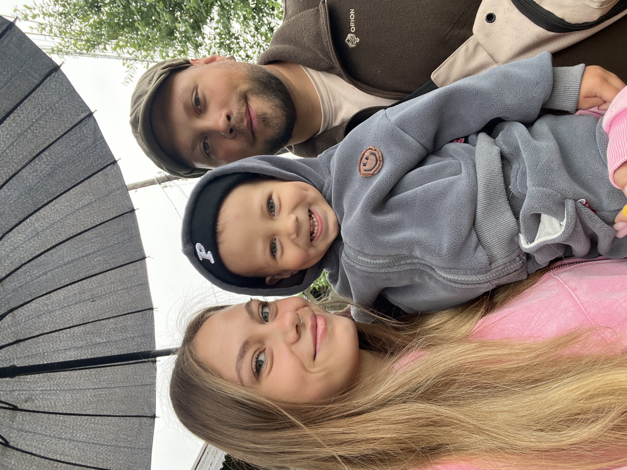 A low-angle, medium shot depicts a family of three. A young woman with long blonde hair and wearing a pink top is at the bottom of the frame. Above her and slightly to the right, a young child with light skin and a grey hooded sweatshirt smiles. Above them is a bearded man wearing a brown jacket and a hat. Part of a black umbrella is visible on the left side of the frame. Tree branches are visible in the upper background. The location appears to be outdoors. There is no information indicating this image was taken in the city of Фоки, Russia.