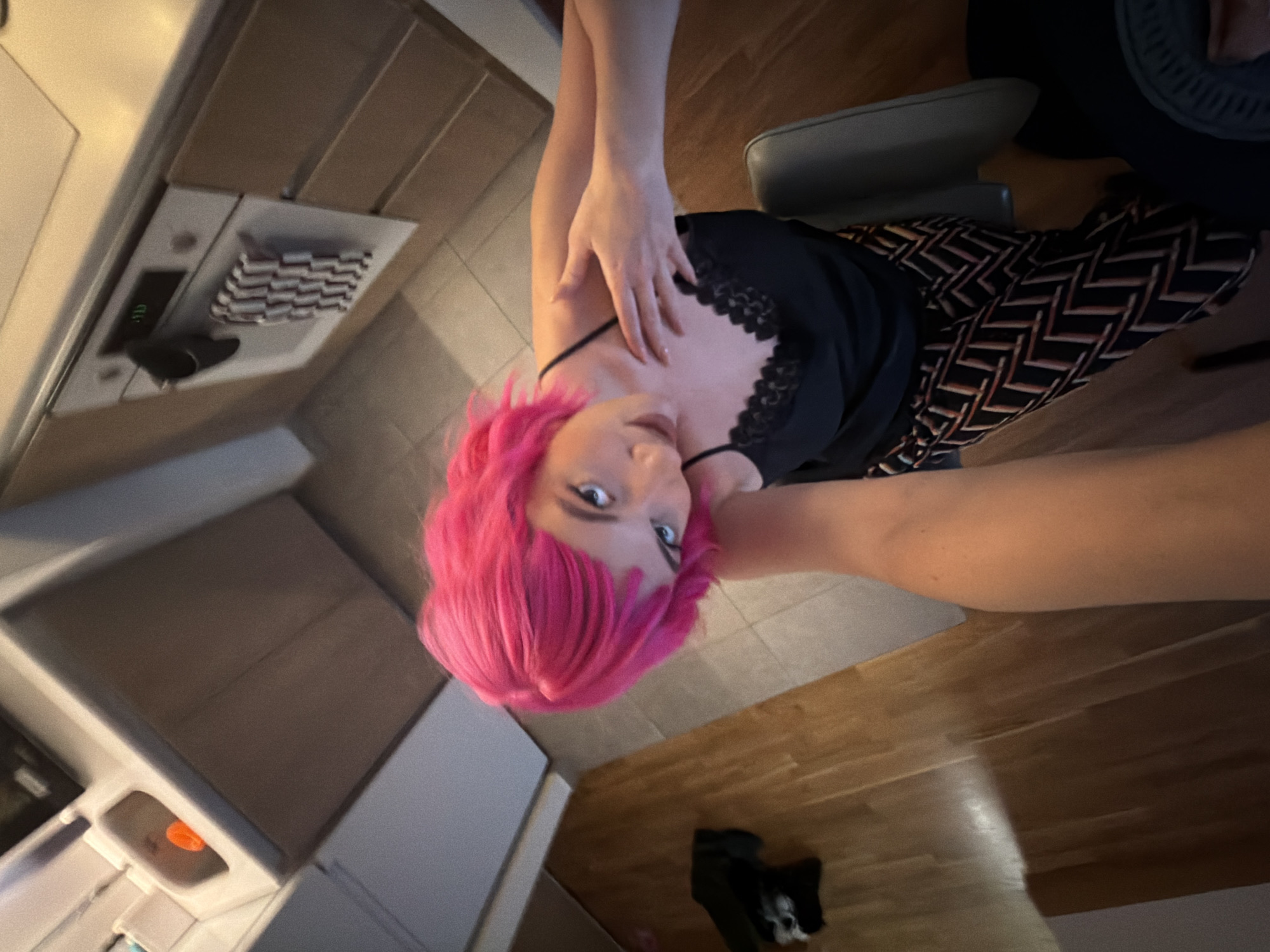 A woman with bright pink hair is pictured in a low-angle, overhead shot. She is wearing a black top with lace detailing and patterned pants. Her right hand is touching her chest. She appears to be indoors, in what may be a kitchen or kitchenette. The scene includes cabinets, a microwave, a dishwasher, a counter, and flooring that transitions from tile to hardwood. A black shoe is on the floor. The location is unknown and there is no evidence that it is in Belgrade, Serbia.