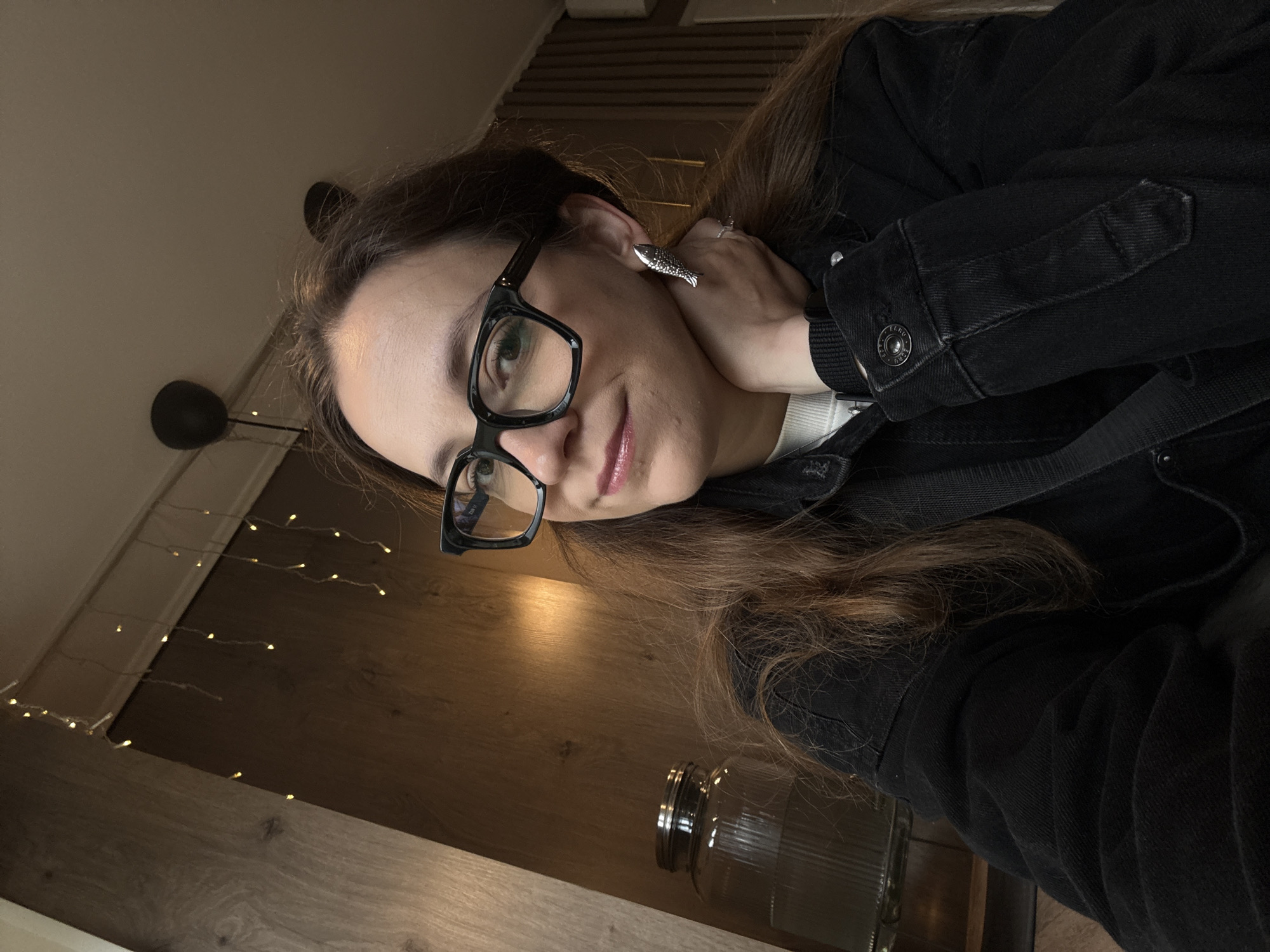 In this close-up, indoor, selfie-style photograph, a woman with long, auburn hair and dark-rimmed glasses poses with her hand resting on her neck. She is wearing a black denim jacket over a white top. A striking, silver fish-shaped earring adorns her left ear. In the background, a large glass jar with a metal lid is visible on a wooden shelf. String lights are draped across the upper part of the background, adding a warm glow to the scene. The overall ambiance suggests a casual and cozy setting.