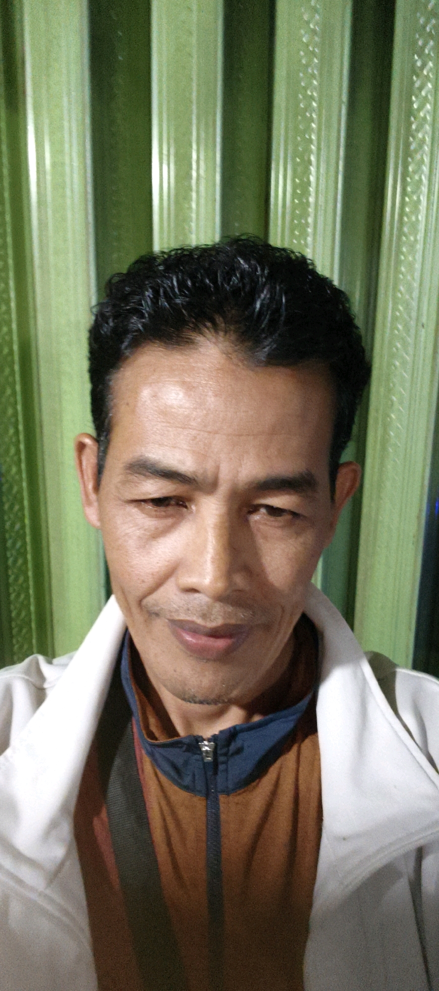 A medium shot portrait of an East Asian man is shown against a green, vertically corrugated background. The man is facing forward, his head tilted slightly down and to his right. He has dark, curly hair, dark eyes, and is wearing a light-colored jacket over a brown shirt with a dark blue collar. A zipper is visible on the front of the brown shirt. His facial expression appears neutral or slightly amused. The lighting is from the front, casting subtle shadows on his face. The background suggests an indoor setting with a decorative wall. The image does not contain any specific details that would allow for pinpointing a location within Muaro Jambi, Indonesia.