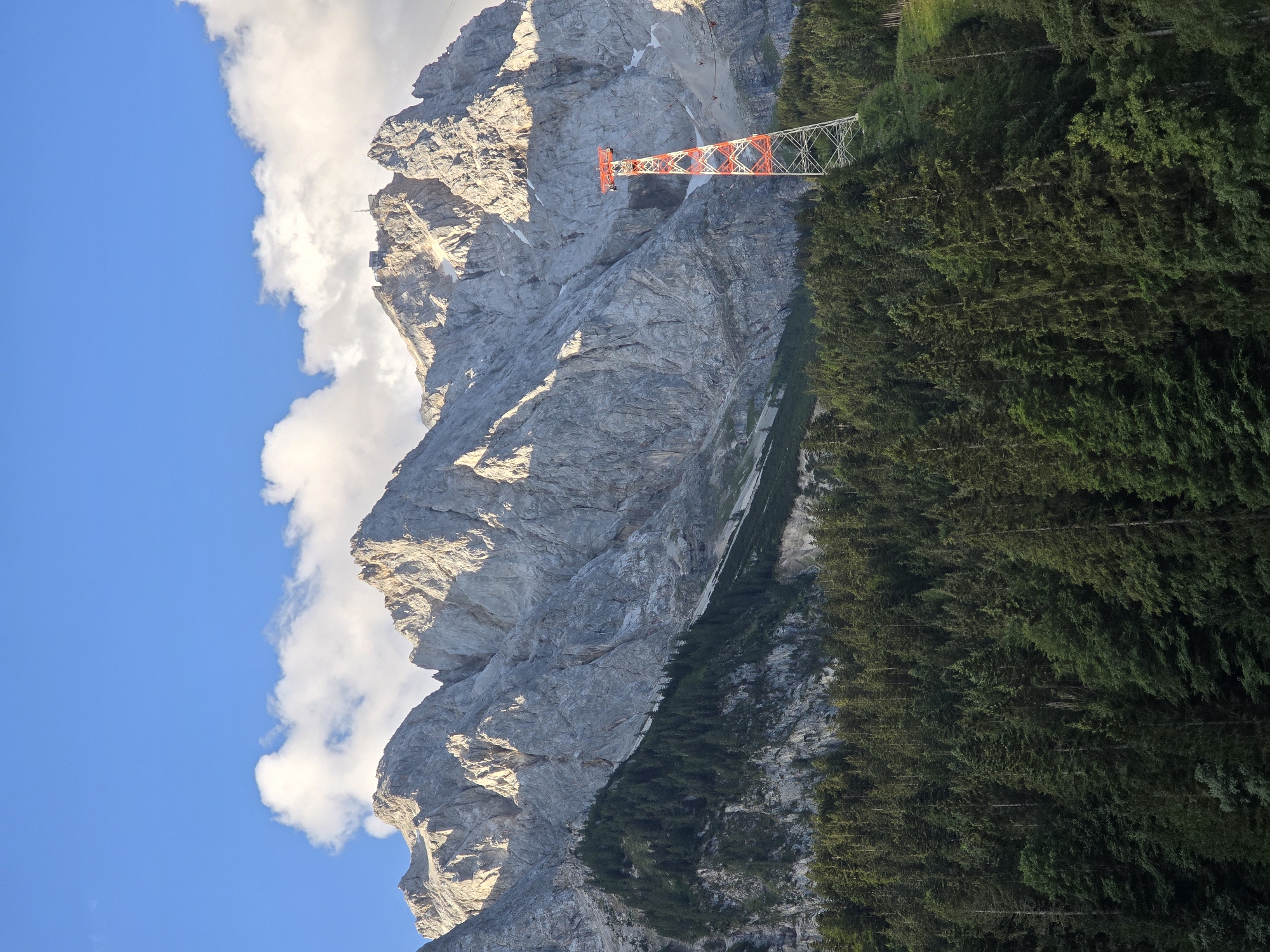 The image depicts a mountainous landscape in Grainau, Germany. In the foreground, a dense forest of dark green coniferous trees covers a sloping hillside. A prominent red and white lattice tower, likely part of a cable car system, stands tall amidst the trees on the right side of the frame. Behind the forest, imposing gray, rocky mountains dominate the background, their peaks casting shadows and highlighting the rugged terrain. The sky above is a vibrant blue, dotted with fluffy white clouds, suggesting fair weather. The lighting indicates it might be late afternoon or early evening, with the sun illuminating the mountain faces.