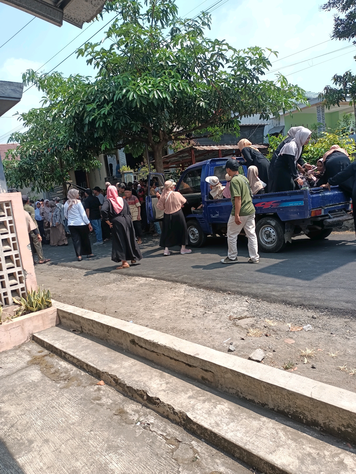 In an outdoor, daytime scene, a group of people, many wearing Islamic head coverings and long robes, are gathered around a blue pickup truck. Some individuals are standing on the ground, while others are seated in the bed of the truck. A man in a green shirt and white pants appears to be assisting or directing the people entering or exiting the truck. The truck is parked on a paved road, with a sidewalk and building visible in the background. Trees with green foliage are also present, suggesting a somewhat vegetated urban or suburban environment. The overall atmosphere appears to be one of a communal gathering or transportation event.