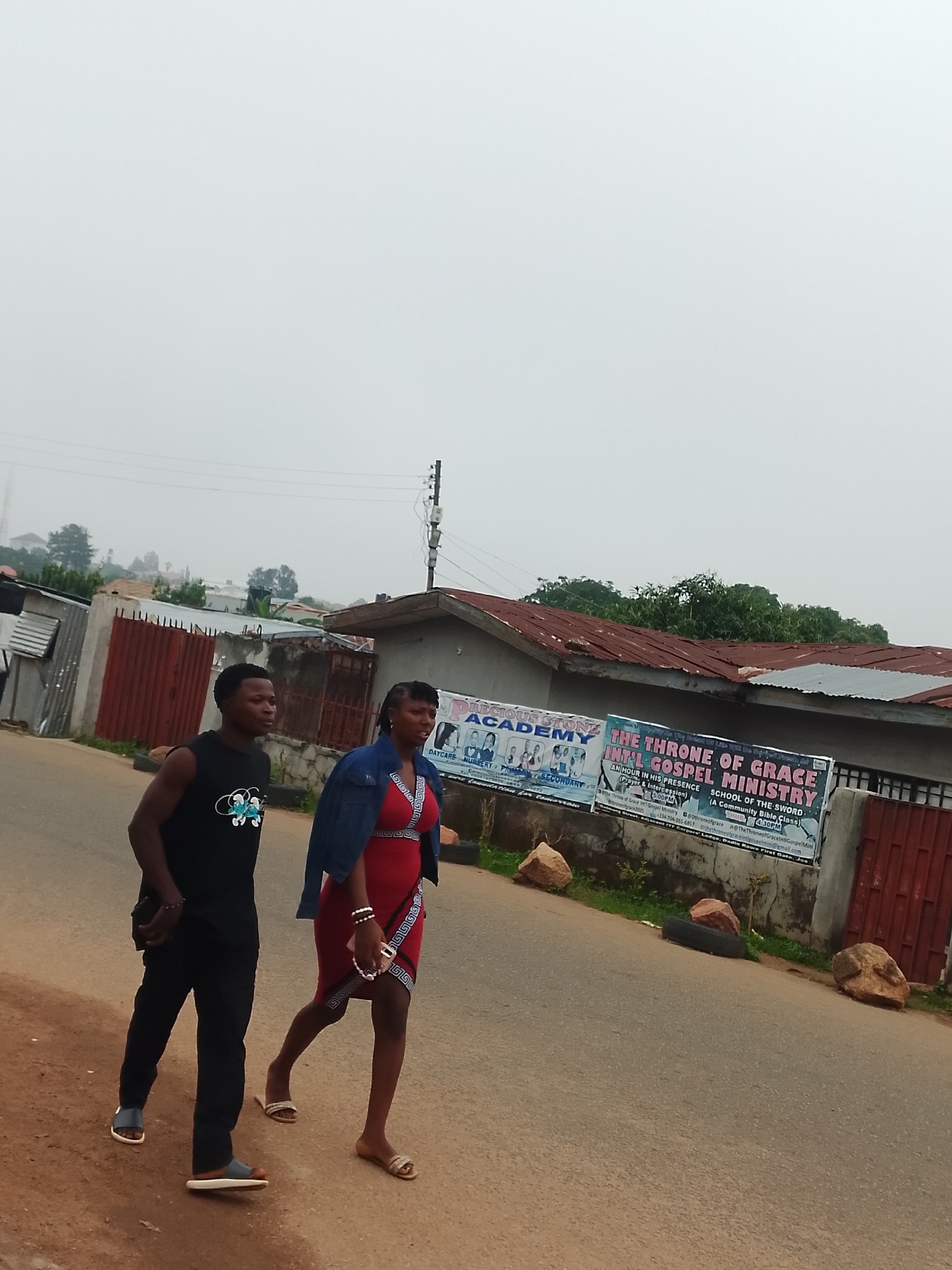 Two individuals, a man and a woman, walk side-by-side on a paved road under an overcast sky. The man, on the left, wears a black sleeveless t-shirt with a graphic print and black pants. The woman, on the right, is dressed in a red knee-length dress and a blue denim jacket. She also wears bracelets on her wrists. Both are wearing sandals. Behind them, on the right side of the road, is a building with a corrugated metal roof and a large banner displaying text and graphics. The banner appears to be advertising an academy and a ministry. The environment is semi-urban, with some buildings and vegetation visible in the background. Scattered on the roadside are several large rocks and a discarded tire.