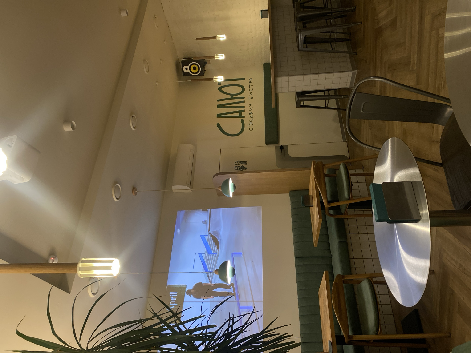 The image shows the interior of a cafe in Krasnoyarsk, Russia, called "Салют Сэндвич Бистро". The cafe appears modern with a minimalist design. There is a row of seating along the wall with wooden chairs and green cushions. Small wooden tables are placed in front of the seating. A taller table made of metal is closer to the camera, with a metal chair. There is a screen showing what appears to be an architectural animation. The name of the cafe is painted on the wall, and a small speaker is above. A potted plant is in the lower left foreground. The floor is made of wood planks.