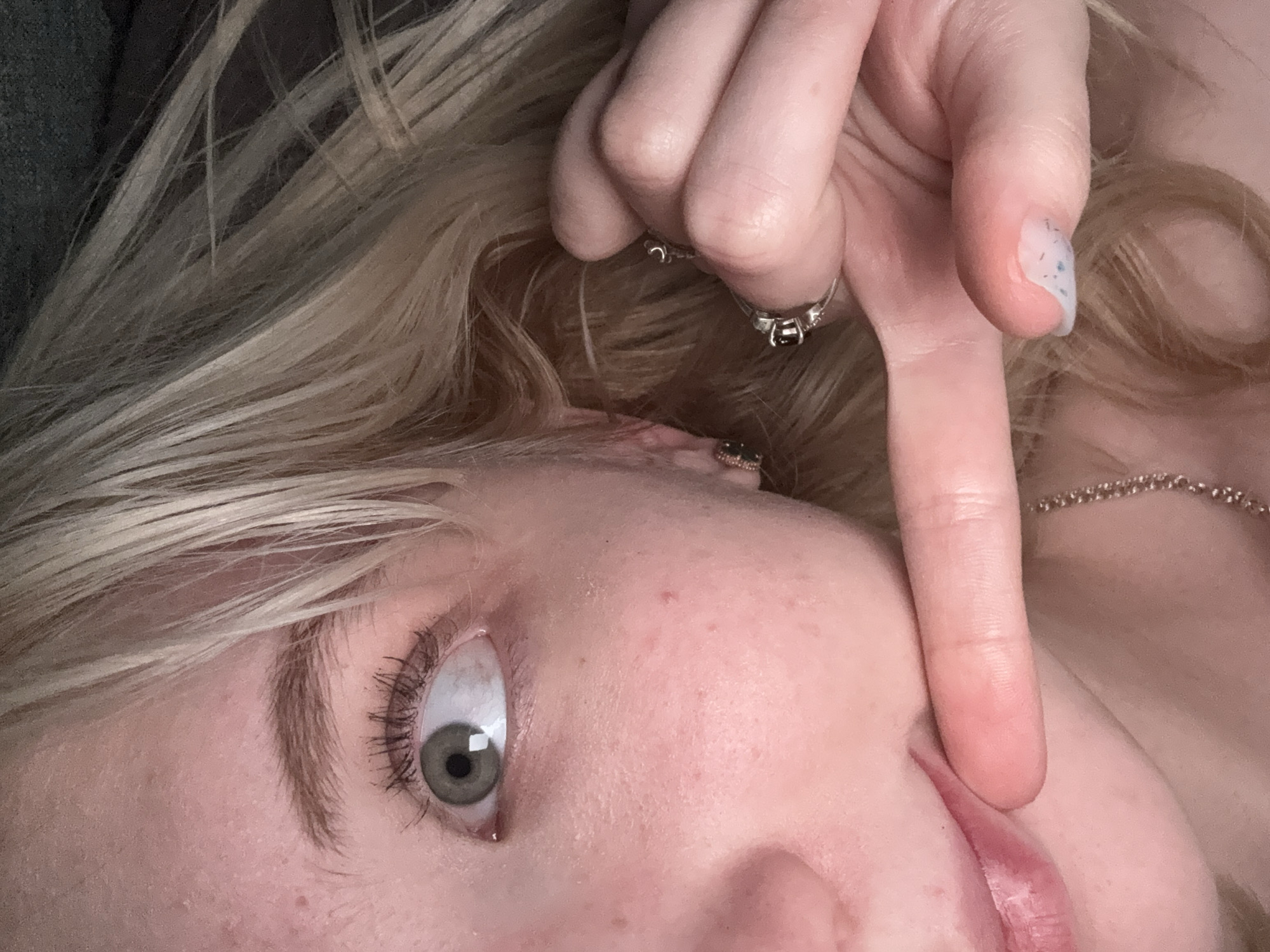 A close-up, low-angle shot shows a portion of a young person's face and hand against a dark background. The person has light skin with some freckles and rosy cheeks. Their left eye, a light green color, is looking to the left, with dark eyelashes and eyebrows. A finger, adorned with two rings and speckled nail polish, is touching the person's lips. A silver necklace is visible around their neck. The lighting is soft and diffused, creating a somewhat moody atmosphere. The background is out of focus, suggesting a shallow depth of field. There are no specific landmarks or environmental cues to indicate a location in a city.