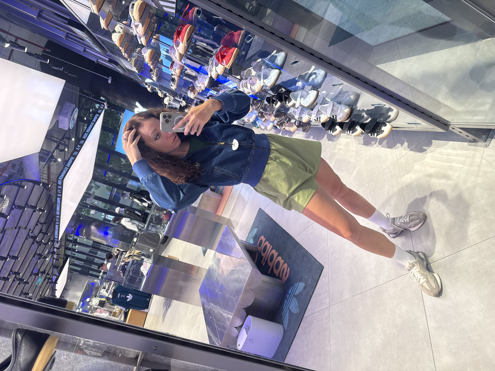 A young woman is taking a selfie in an Adidas store, as seen in the reflection of a mirrored wall. The store is located in Hà Nội, Vietnam. The woman is dressed in a denim jacket, a green dress or skirt, and white socks with grey sneakers. She is holding a smartphone up to take the picture, and her hand is touching her hair. The store has shelves filled with Adidas shoes. In the foreground, a display unit and a rug with the Adidas logo are visible.