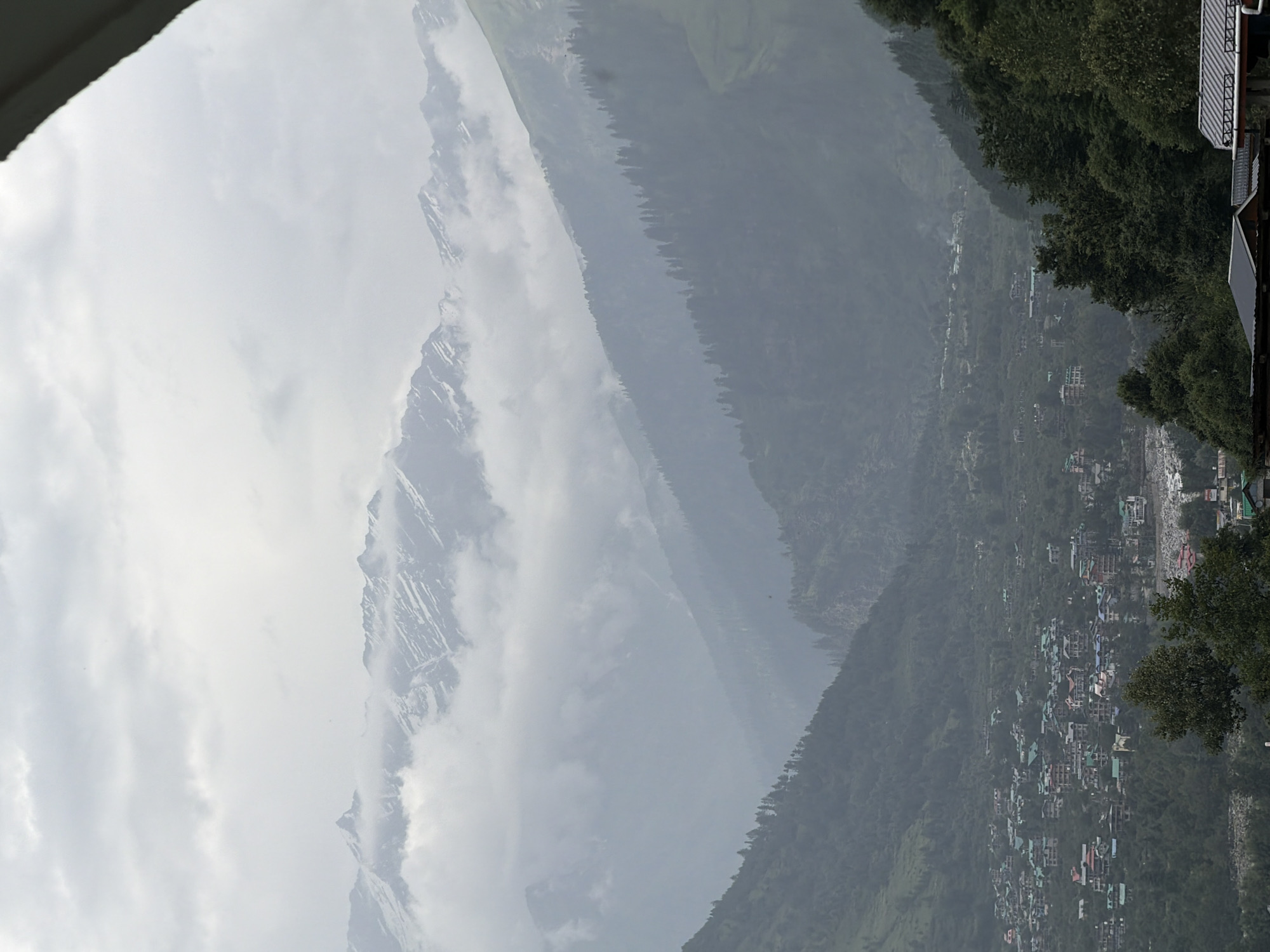 The image depicts a panoramic view of the village of Vashisht, India, nestled within a valley of lush green mountains. In the foreground, a cluster of houses with colorful roofs, characteristic of the region, are visible, interspersed with dense vegetation and trees. A river can be seen winding its way through the valley, adding to the scenic beauty. The midground is dominated by steep, tree-covered slopes that rise dramatically towards the sky. Further in the background, majestic snow-capped mountains are partially obscured by a thick layer of clouds, creating a dramatic and ethereal atmosphere. The sky is overcast, with a muted light that lends a soft, diffused quality to the scene. There are no visible people in the image. The overall impression is one of a tranquil and picturesque mountain village.