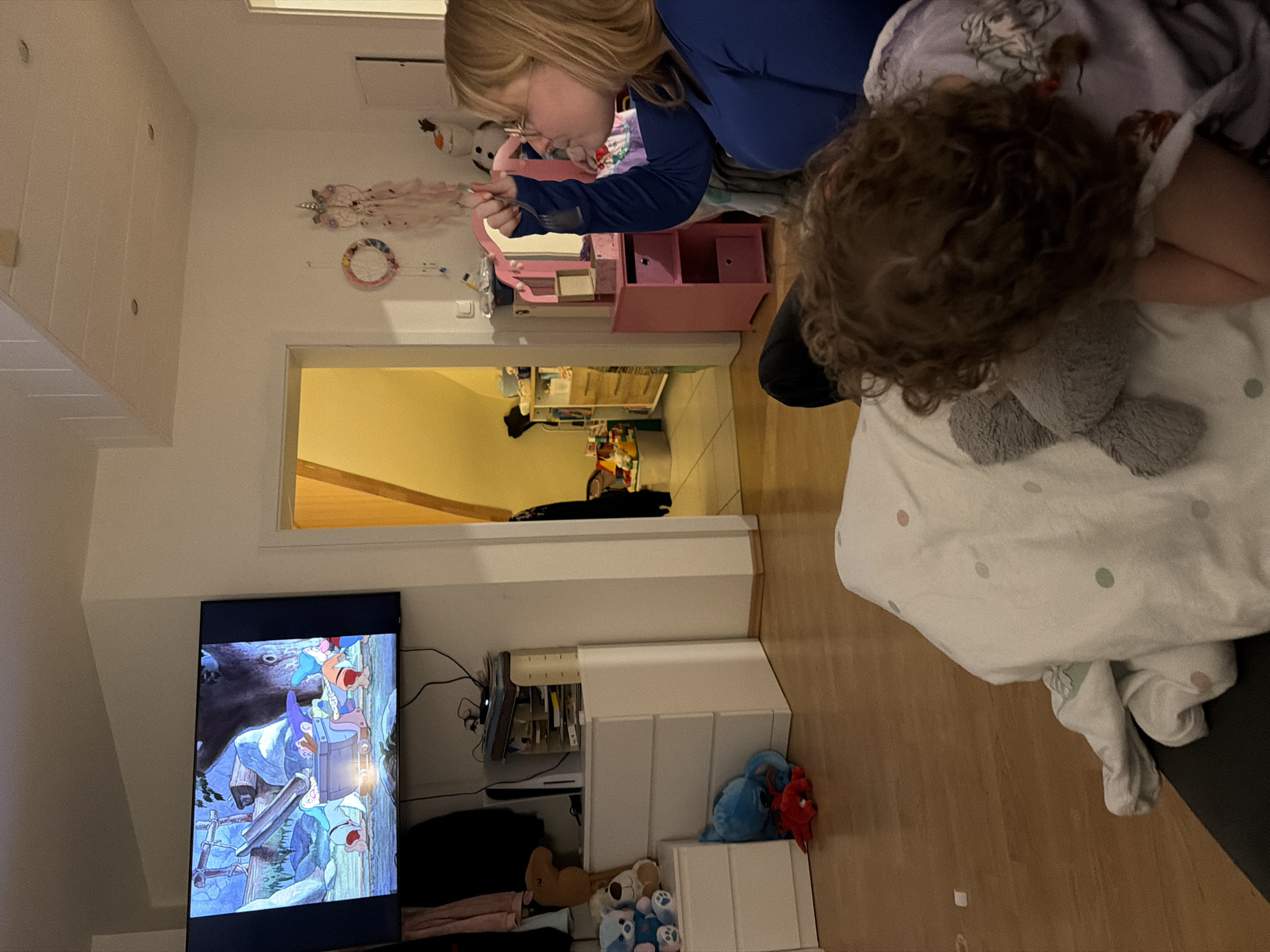 The image depicts an indoor scene, likely a living room or bedroom, in Trassem, Germany, centered around two individuals relaxing. On the left, a large television screen displays an animated scene, possibly from "Snow White and the Seven Dwarfs," featuring several dwarves in a mine.
In the foreground, on the right, a person with curly brown hair rests on a white duvet with light pink and green polka dots, cradling a grey plush toy. To their left, a blonde-haired individual, wearing glasses and a blue long-sleeved top, is seen in profile, holding a fork or spoon, suggesting they are eating.
The room features light wooden flooring and white walls. Behind the blonde individual, a pink vanity table or desk is visible, along with various wall decorations including a white plush Olaf doll, a pink feathered dream catcher, and colorful circular ornaments. A doorway in the mid-ground opens into another room or hallway, illuminated with a warm, yellowish light, possibly revealing a staircase.
Below the television, a white modular shelving unit holds several items, including cables, a book holder, and a collection of plush toys such as blue and red creatures, a brown teddy bear, and smaller blue bears. The lighting suggests a relaxed evening atmosphere in a comfortable, family-oriented home. No discernible text is visible within the frame.