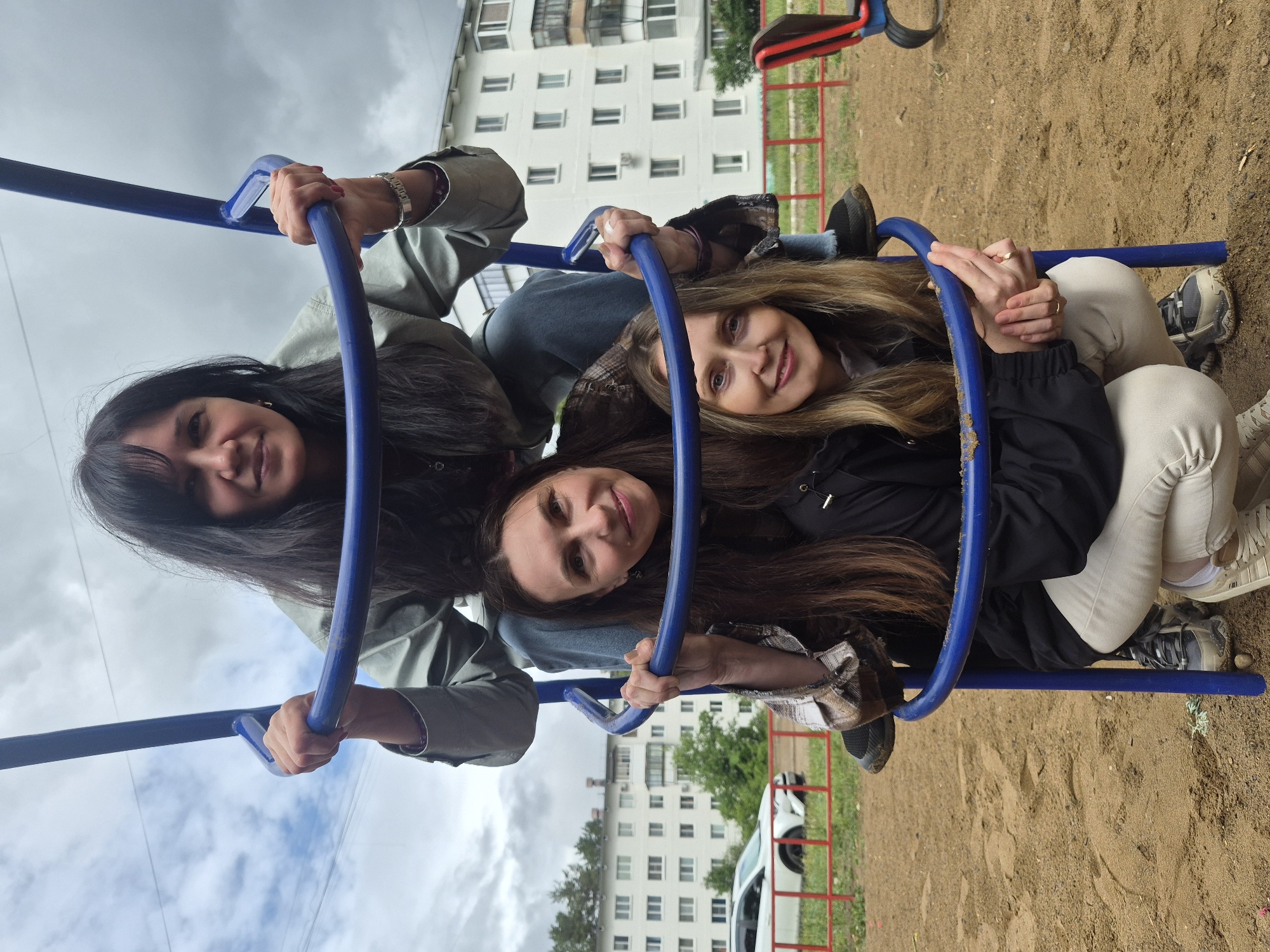 Three women are crammed into a blue playground climbing frame. The woman on the left has long, dark hair and is wearing a green jacket. The woman in the middle has long, brown hair and is wearing a denim jacket. The woman on the right has long, blonde hair and is wearing a black jacket and beige pants. They are all smiling. The location appears to be a playground in an urban area. In the background, there is a multi-story building with white walls and many windows. The ground is covered in sand. There is a white car parked near a red fence in the background.
