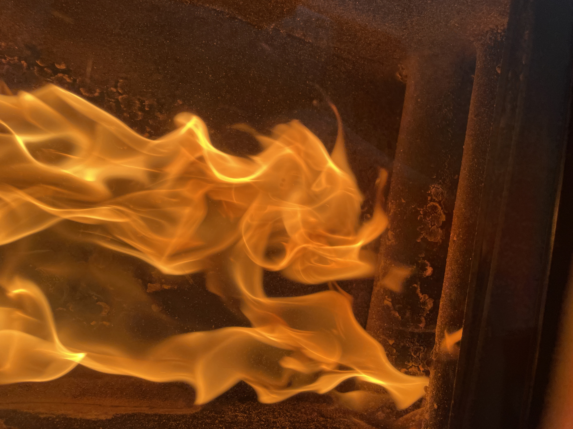 The image depicts flames within a confined, possibly metallic, structure. The flames are predominantly orange and yellow, exhibiting dynamic movement and swirling patterns. To the right are three vertical cylindrical objects, possibly pipes, with a dark, textured surface. Above the flames is a dark, grainy background that could be a wall or the interior of the structure. No people are visible. The location is not identifiable as Zlatograd, Bulgaria based on the visual information.