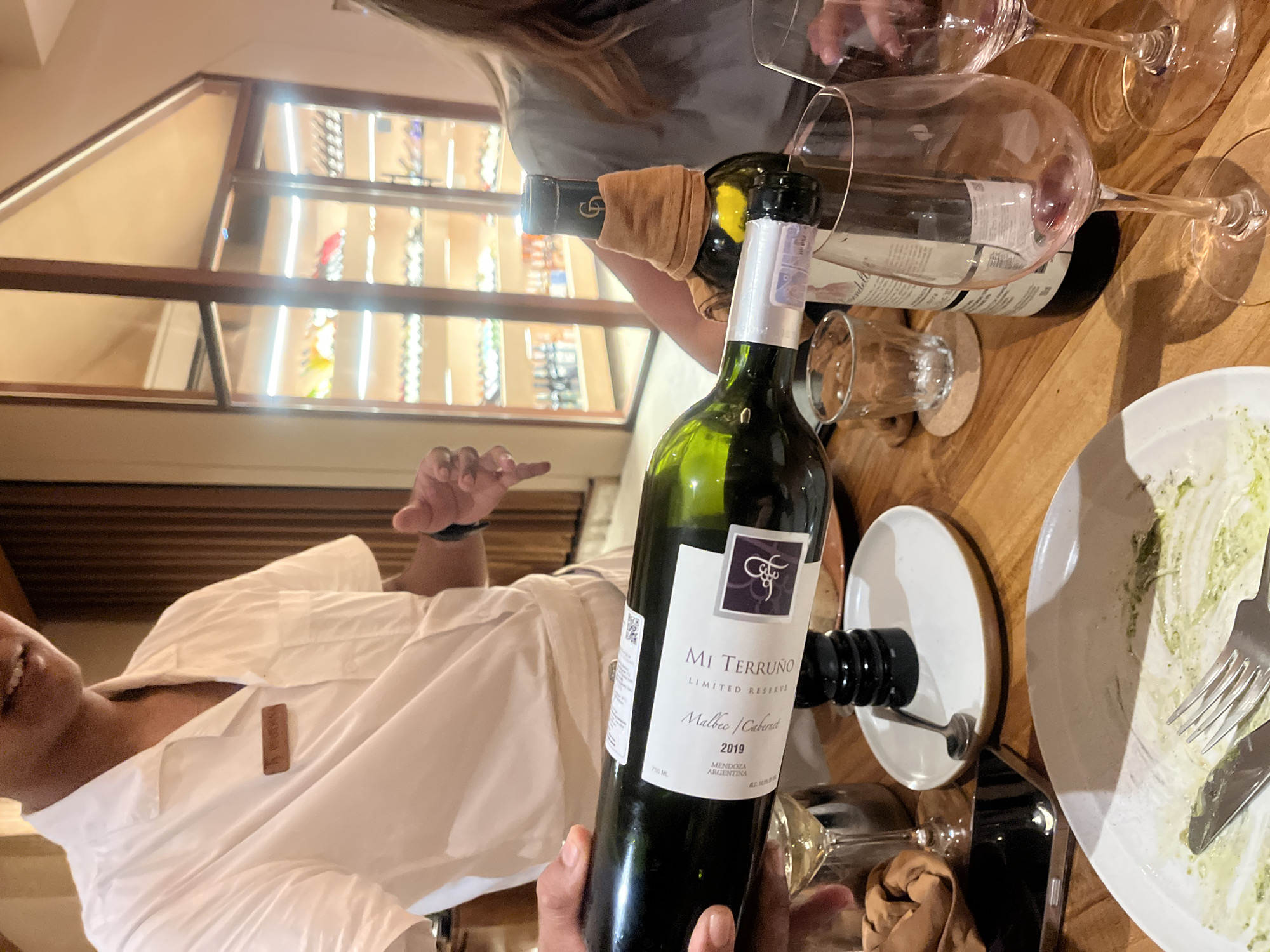 A server in a white shirt presents a bottle of Mi Terruño Limited Reserve Malbec/Cabernet 2019 wine at a table set with wine glasses, small drinking glasses, plates, and silverware. The background shows a lighted display of wine bottles. The location may be in Kuta Selatan, Indonesia.