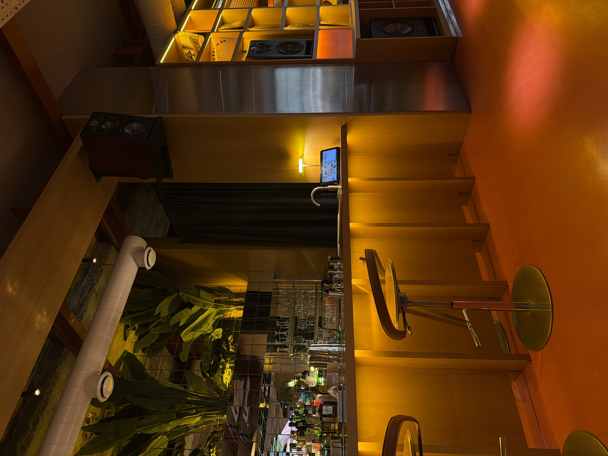 The image depicts a bar setting in Yerevan, Armenia. The interior features a multi-tiered shelf displaying various objects, including speakers and a book labeled "FONOTECA." Below the shelf is a countertop with a small screen mounted above a sink. A bar stool is positioned in front of the counter. Behind the counter, there's a green curtain or partition. A mirrored section reflects the bar's interior, showcasing glasses, bottles, and plants. A white duct or pipe runs vertically along the left side of the mirrored section. The color palette includes warm tones like yellow and orange, with wooden and metallic elements throughout. The scene appears unoccupied, focusing on the architectural and design aspects of the bar.