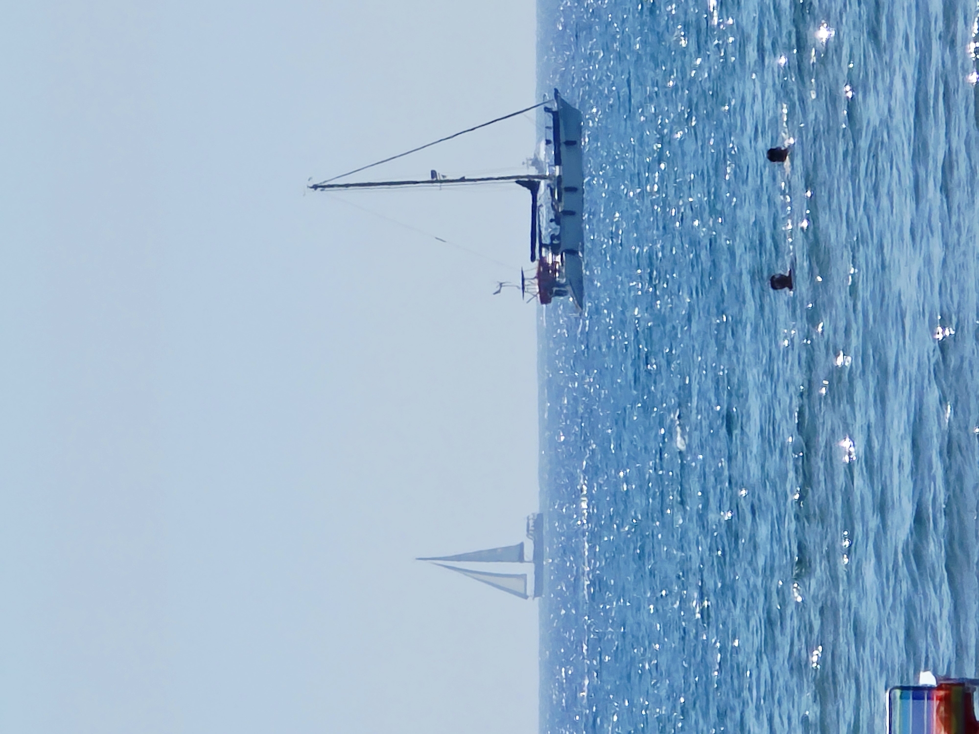 Two sailboats are visible on a bright, clear day, with the closer sailboat appearing larger and more detailed. The water is a vibrant blue with sparkling reflections from the sun. In the foreground, two people are swimming in the sea, with only their heads and shoulders visible above the water. A portion of a colorful beach chair is visible in the lower left corner. The scene suggests a leisurely day at the beach or sea, likely in a coastal area such as the coastline of Larnaca, Cyprus, known for its blue flag beaches and clear waters.