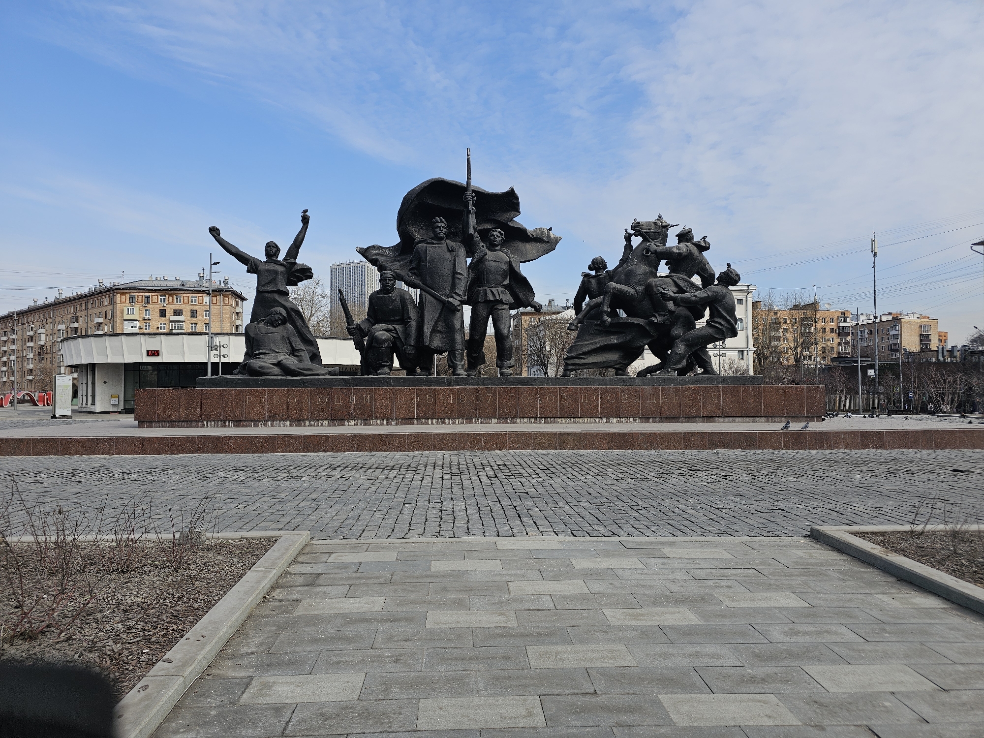 This image captures the "Monument to the Fighters of the 1905 Revolution" on Krasnaya Presnya Street in Moscow, Russia.
The monumental sculpture, rendered in dark bronze, occupies the central midground. It is composed of multiple figures depicting scenes from the 1905-1907 revolution. On the left, a female figure stands with arms raised, a seated figure below her. In the center, a group of standing and kneeling male figures, one prominent figure holding a flag pole, are positioned against a large, flowing flag. On the right, a dynamic equestrian group features at least two figures on horseback, engaged in struggle with other figures on the ground. Several pigeons are perched on the right-hand sculptural elements. The entire monument rests on a multi-tiered base of reddish-brown polished stone, featuring the inscription "РЕВОЛЮЦИИ 1905-1907 ГОДОВ ПОСВЯЩАЕТСЯ" (Dedicated to the Revolution of 1905-1907) in Cyrillic lettering.
The foreground consists of light gray rectangular paving slabs, transitioning to a darker gray cobblestone-patterned pavement. A narrow strip of barren earth with sparse, dormant shrubbery separates these two paved sections.
In the background, under a bright blue sky with scattered white clouds, a cityscape is visible. To the left, a multi-story building with a light-colored ground floor and upper brick-colored sections features a digital clock displaying "11:42." Further back, various residential and commercial buildings are seen, including a distant tall, modern white skyscraper. Overhead, utility lines and poles, including a cellular antenna, crisscross the sky. Leafless trees are interspersed throughout the urban landscape. A few pigeons are also visible on the cobblestone pavement near the monument's base.