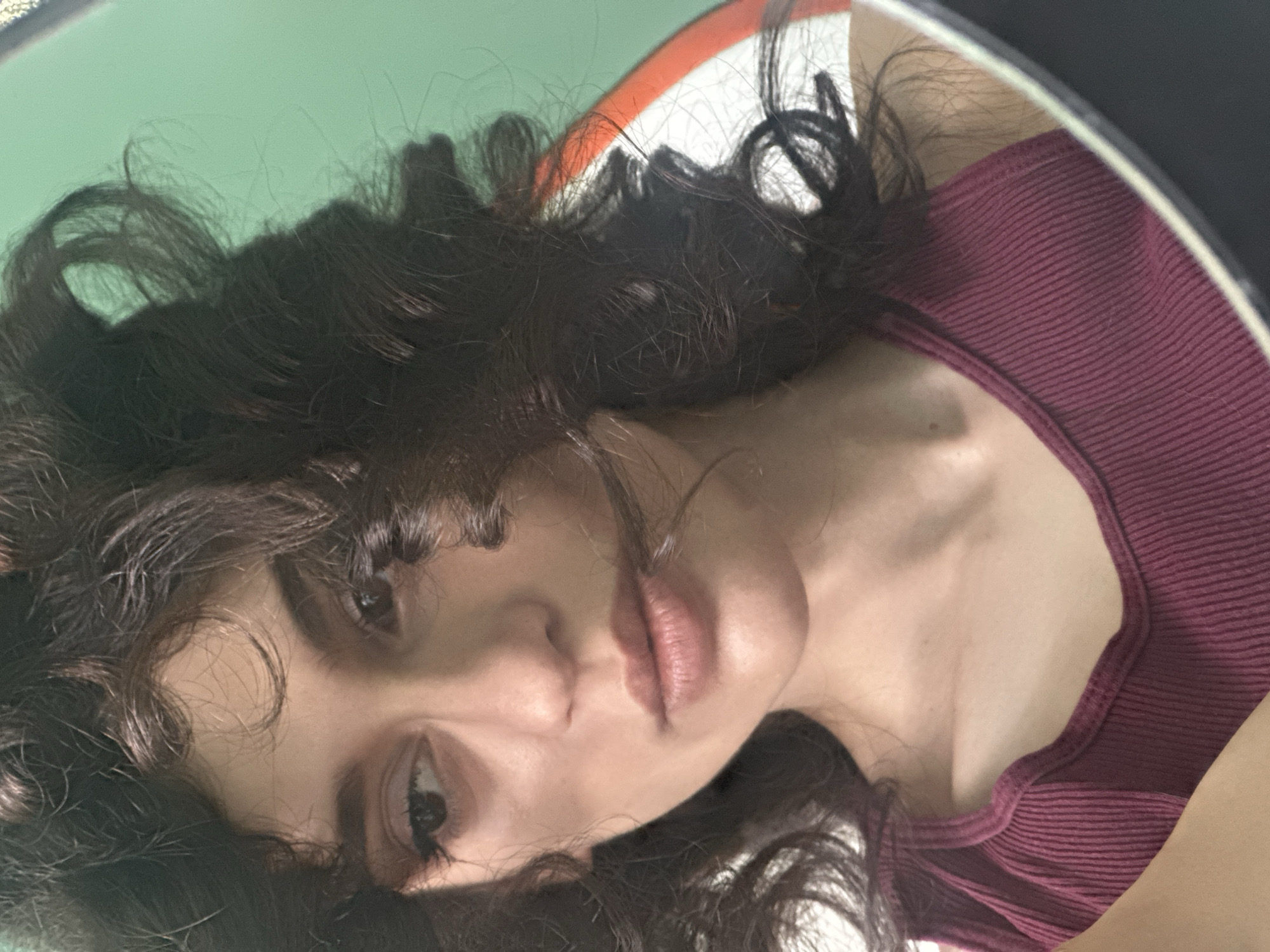 The image is a close-up, reflected shot of a young woman with dark, curly hair and fair skin, looking to her left. She is wearing a burgundy ribbed tank top. The background is a plain light green wall, with a partial view of a circular mirror frame featuring white and orange segments. The woman's facial features are visible, including her eyes, nose, and lips. The setting appears to be indoors, with no specific identifiable landmarks of Sofia, Bulgaria.