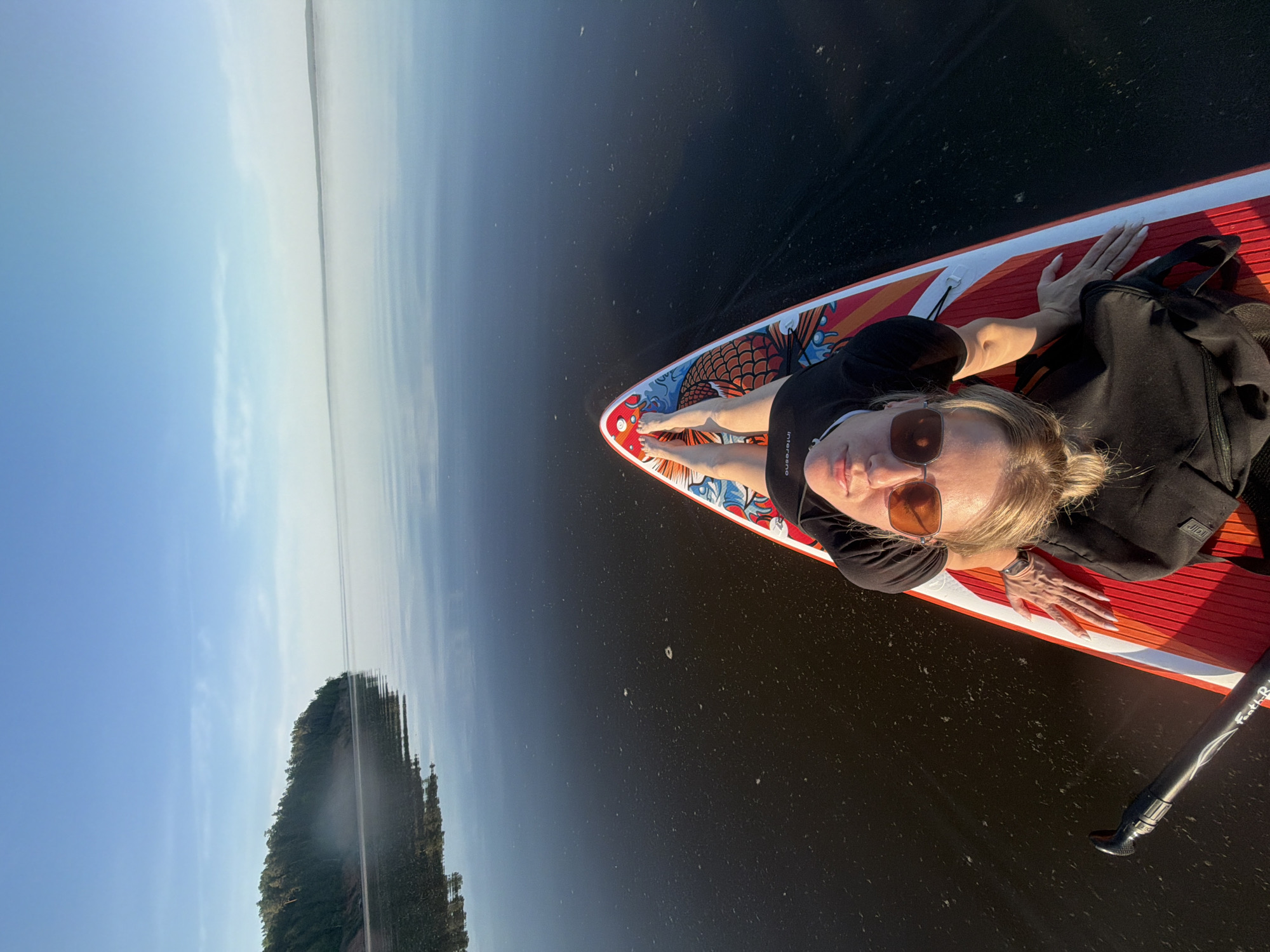 A woman with sunglasses is lying on a red paddleboard on calm water. The paddleboard has a koi fish design on the front. The water reflects the sky, which transitions from blue to a light, hazy white. A forested landmass is visible in the distance on the left side of the image. A paddle lies alongside the board. The location is likely a body of water near Noviy, Russia, inferred from the image's caption.