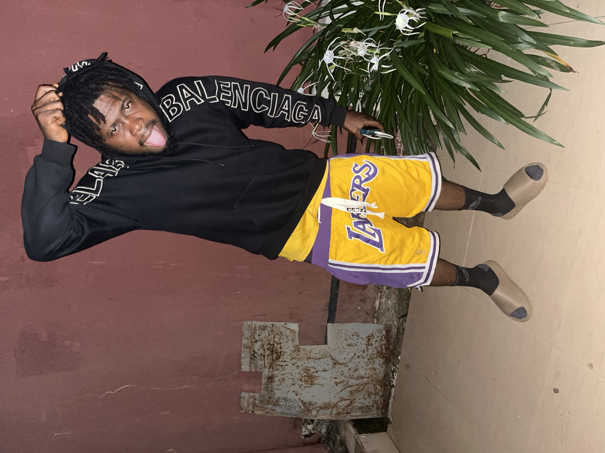 The image shows a young man standing in front of a reddish-brown wall. He is wearing a black hoodie with white lettering on the sleeves and a pair of yellow basketball shorts with purple trim and a Lakers logo. He has black socks and tan slides on his feet. The man is sticking his tongue out and pulling on the hood of his sweatshirt. To his right is a large green plant with white flowers. To his left is a rusted metal object against the wall, and the ground is tiled. The scene appears to be outdoors, possibly in an alleyway or backyard. There is no specific indication of the city of Jos, Nigeria, within the image.