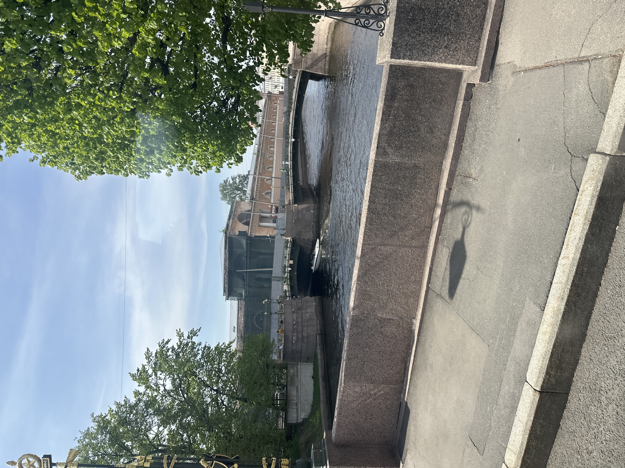 The image depicts a canal in Saint Petersburg, Russia. The vantage point is from a sidewalk alongside the canal, with a granite retaining wall on the right. The canal is narrow, with buildings visible on the opposite bank. The architecture on the far bank includes arched structures and building elements under renovation. A small boat is in the water. Trees line the canal banks. The sky is blue with some clouds. The sidewalk shows cracks and a shadow of a lamp post.