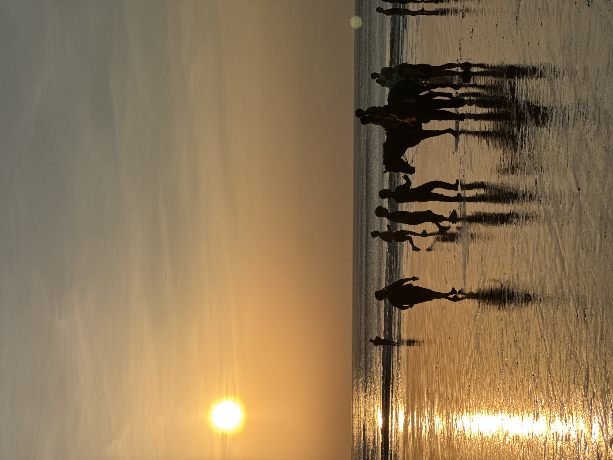 The image depicts a beach scene at sunset in Kuta Selatan, Indonesia. Silhouettes of people are visible on the wet sand, with some individuals running and others standing. A horse with riders is also present, with several people gathered around it. The sun, a bright orb, is low on the horizon, casting a warm golden glow across the sky and reflecting on the water and wet sand. The ocean is calm with gentle waves. The overall impression is of a peaceful evening at the beach.