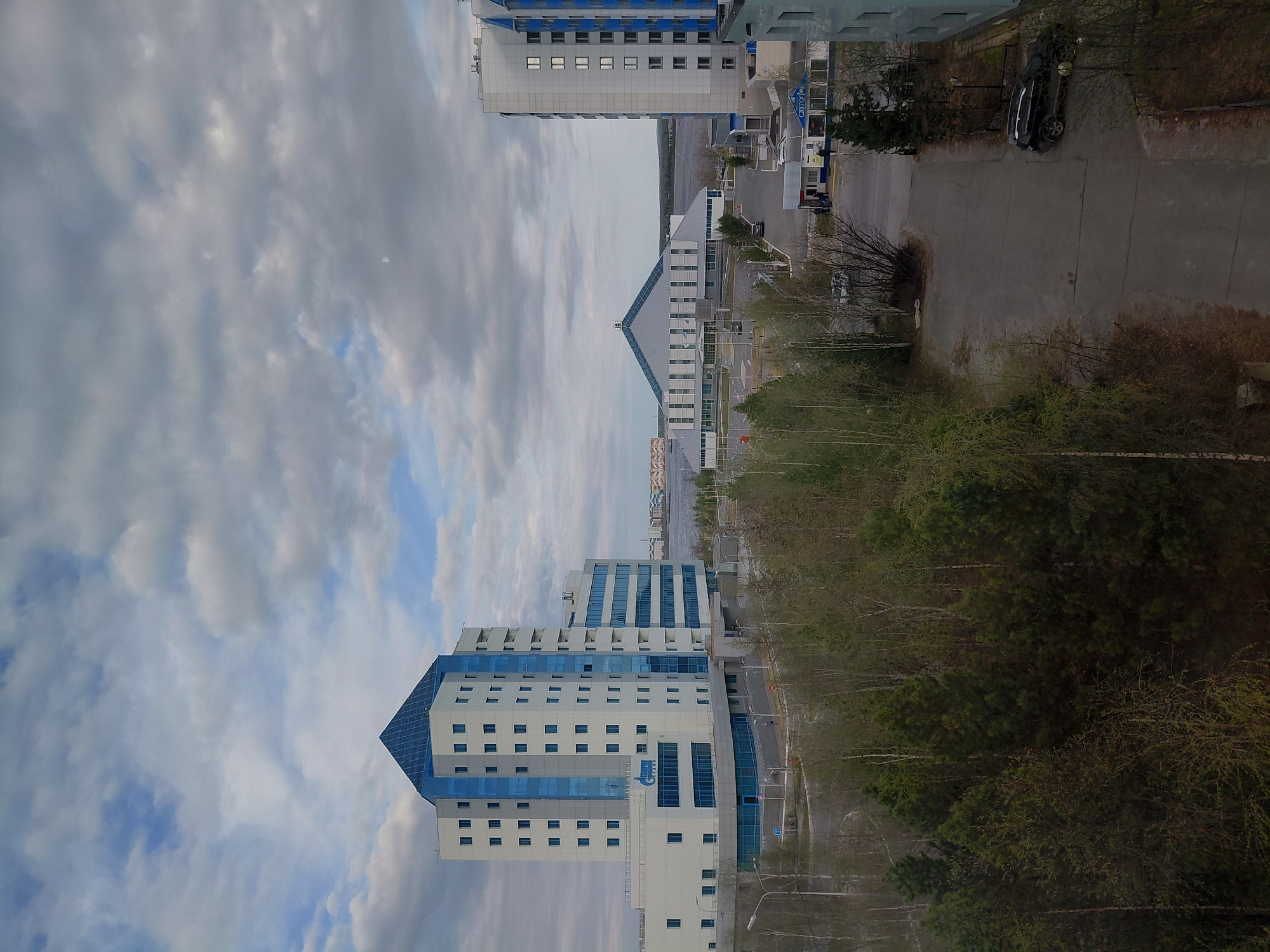 The image shows a cityscape in Nadym, Russia, with a view of two prominent buildings. The first building, located in the foreground, is a multi-story structure with a white facade, blue glass panels, and a distinctive blue, pyramid-shaped top. The "Gazprom" logo is visible on one of its sides. Behind it, there's another building, white with a triangular, pitched roof, and a larger, less distinctive building further back. A road runs between the buildings, with a few parked vehicles and some vegetation along the edges. Trees are heavily populated around the buildings. The sky is partly cloudy with a mixture of gray and white clouds. The scene appears to be captured from an elevated vantage point, providing an overview of the buildings, road, and surrounding environment.