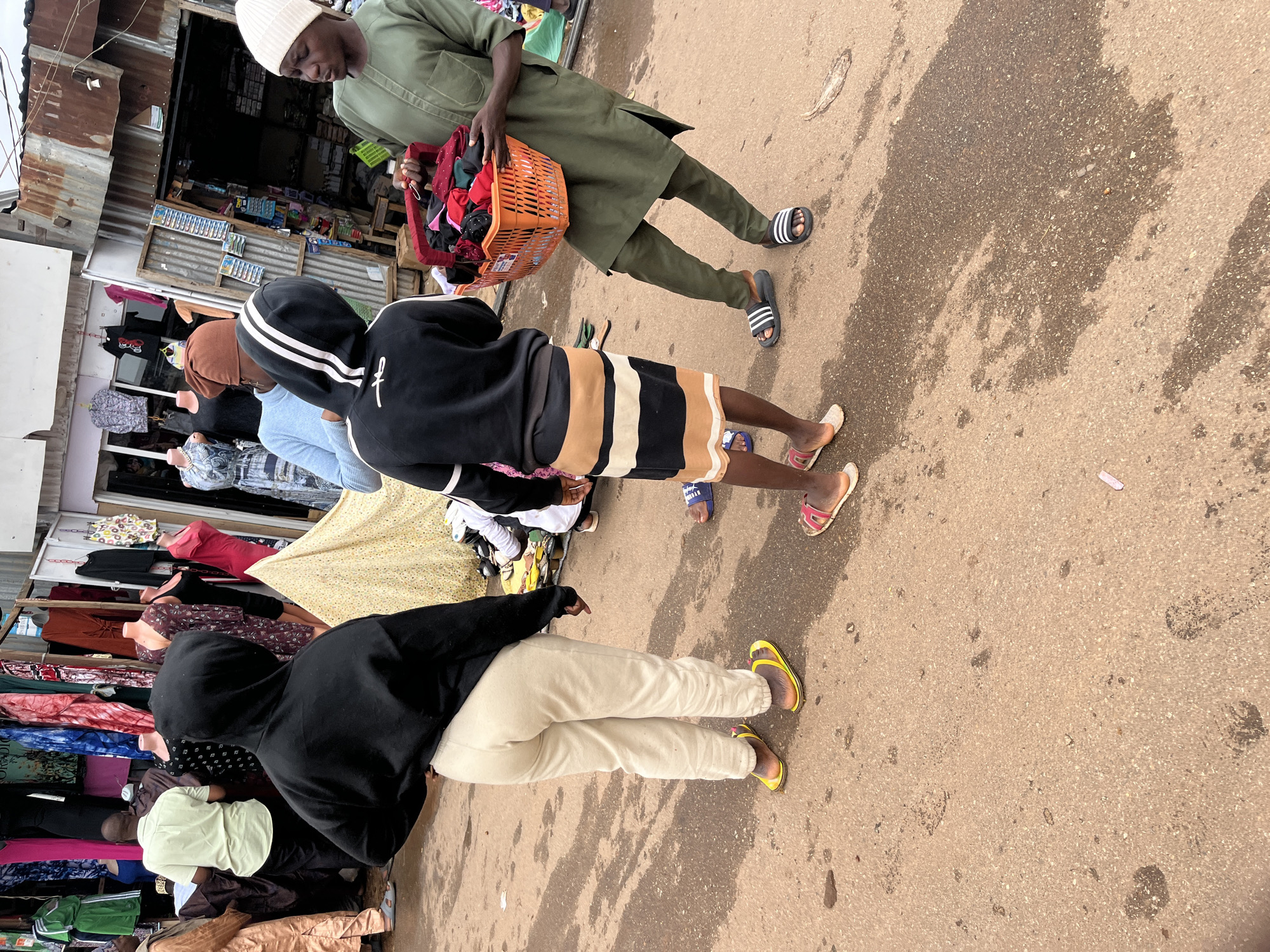 This is a full-shot, eye-level, outdoor photograph of a street market in Jos, Nigeria. The ground is a dirt or asphalt mixture with some wet patches and debris. In the foreground, three individuals are visible from the waist down, engaged in interaction.
To the left, a person wearing a black hooded sweatshirt and light beige pants stands facing away from the camera, with their left hand on their hip and their right arm bent and slightly raised. They are wearing bright yellow flip-flops.
In the center, a younger person wearing a black hooded sweatshirt with white stripes on the sleeves and a striped skirt is also facing away, looking towards the right. They are wearing pink and white flip-flops.
To the right, a person wearing a green traditional outfit and a white beanie is holding an orange woven basket filled with clothing items. They are facing the younger person in the center and appear to be engaged in conversation or a transaction. This person is wearing black slides with white stripes.
In the background, market stalls are visible with various clothing items displayed on mannequins and racks. The stalls are constructed with corrugated metal and wood. A patterned yellow tent is also visible behind the central figure. The overall scene depicts a lively commercial environment.