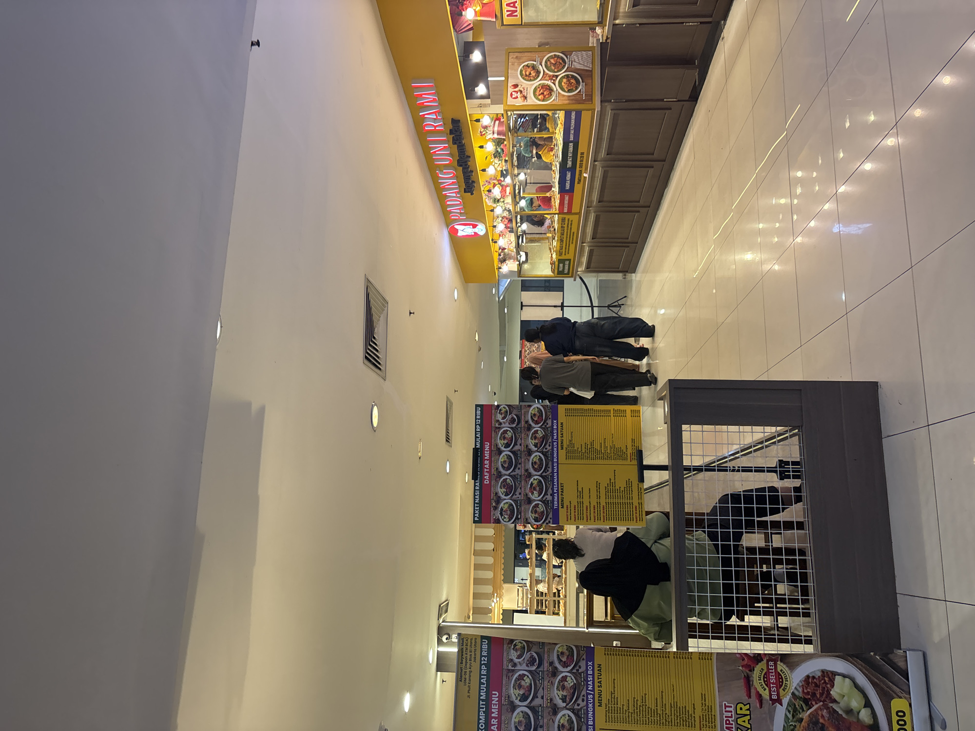 The image shows a food court in a mall, likely in North Jakarta, Indonesia. A prominent yellow sign reads "PADANG UNI RAMI" with "Kue Kering & Aneka Takjil" below it, indicating a Padang cuisine restaurant. To the left, a menu board for "NASI BUNGKUS / NASI BOX" is visible, with "PAKET LENGKAP MULAI RP 12 RIBU" printed on it. Another menu board to the right displays "PAKET NASI RAMES MULAI RP 12 RIBU". Several food stalls are arranged along a counter, with staff members preparing food. In the foreground, two individuals are seated at a table, one wearing a green hijab. Other patrons are walking through the food court. The floor is tiled and reflects the overhead lighting.