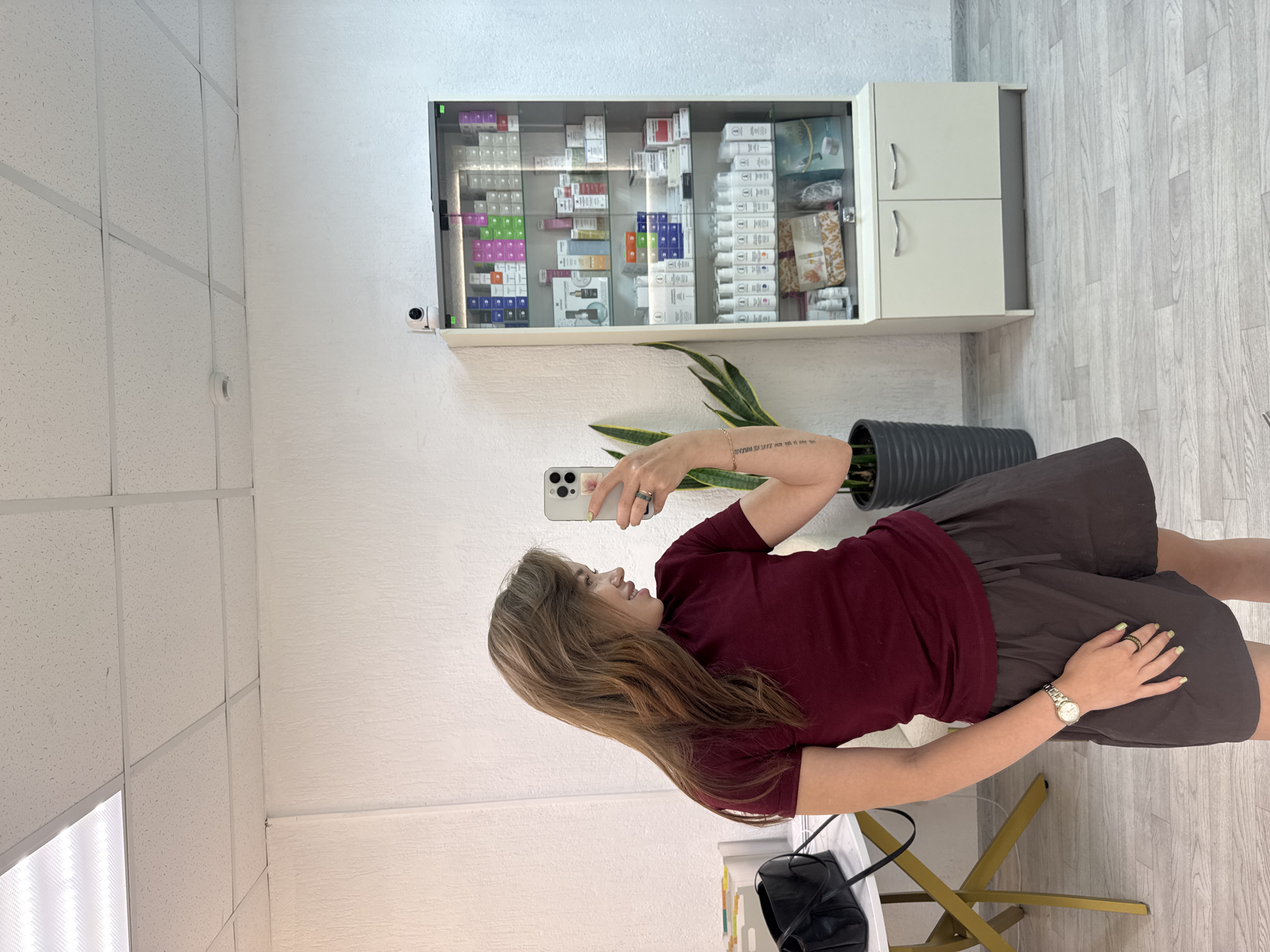 A woman is taking a selfie in a room. She is wearing a maroon t-shirt and dark grey shorts. She has long, brown hair and is wearing a watch on her left wrist and a ring on her right index finger. In the background, there is a display cabinet filled with various bottles and boxes, a plant in a pot, a table with a black bag, and a security camera mounted on the wall. The room has white walls and a white tiled ceiling. The floor is light-colored wood. The woman is smiling. This is an indoor scene, likely a retail or service establishment, located in the city of Chaikovsky, Russia.