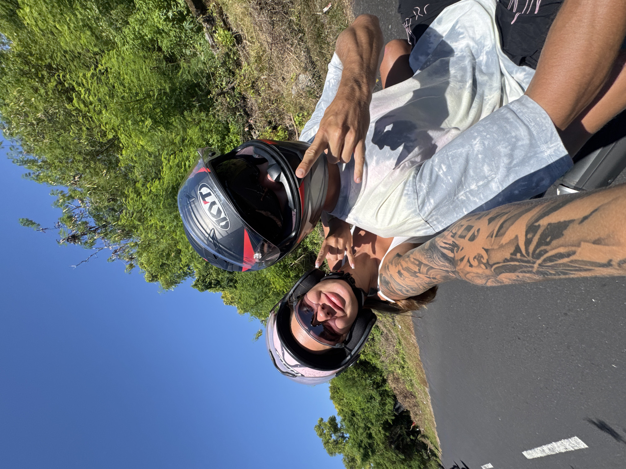 A selfie captures two individuals on a motorcycle, likely traveling along a road in Denpasar, Indonesia, as inferred from the environment. Both are wearing helmets. The person in the foreground has sunglasses on. The other person is holding up the "rock on" hand gesture. Lush green foliage lines the road, contrasting with the clear blue sky.