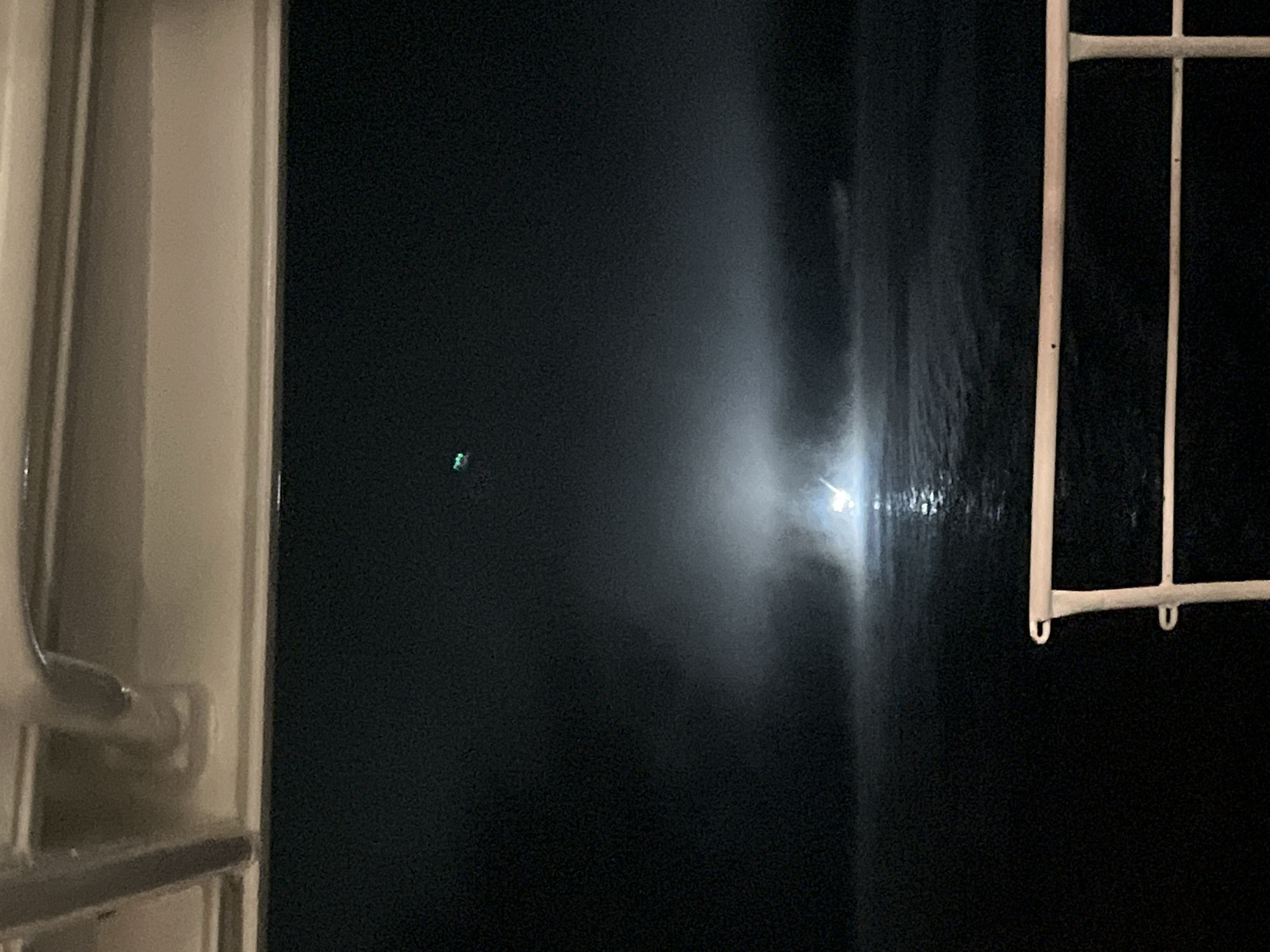 The image shows a dark, possibly foggy or misty night scene over water. In the distance, a bright light source illuminates the fog, creating a dramatic glow. A small green light is visible in the upper portion of the sky. The foreground features a white railing, suggesting the viewpoint is from a boat or a structure over the water. The overall atmosphere is somber and mysterious. The location in Novoilinsky, Russia, is not visually evident from the provided image.